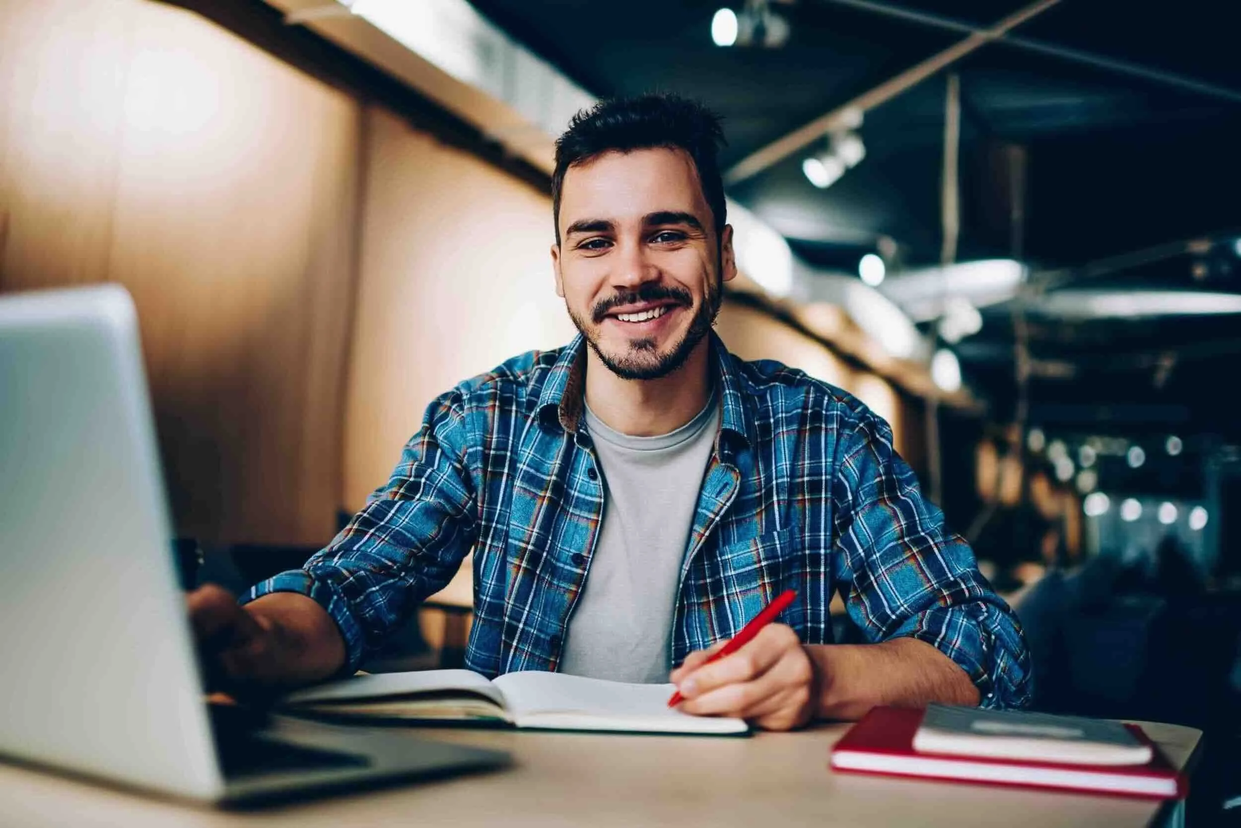 Student applicant in a plaid shirt smiles while studying at a desk with a laptop.