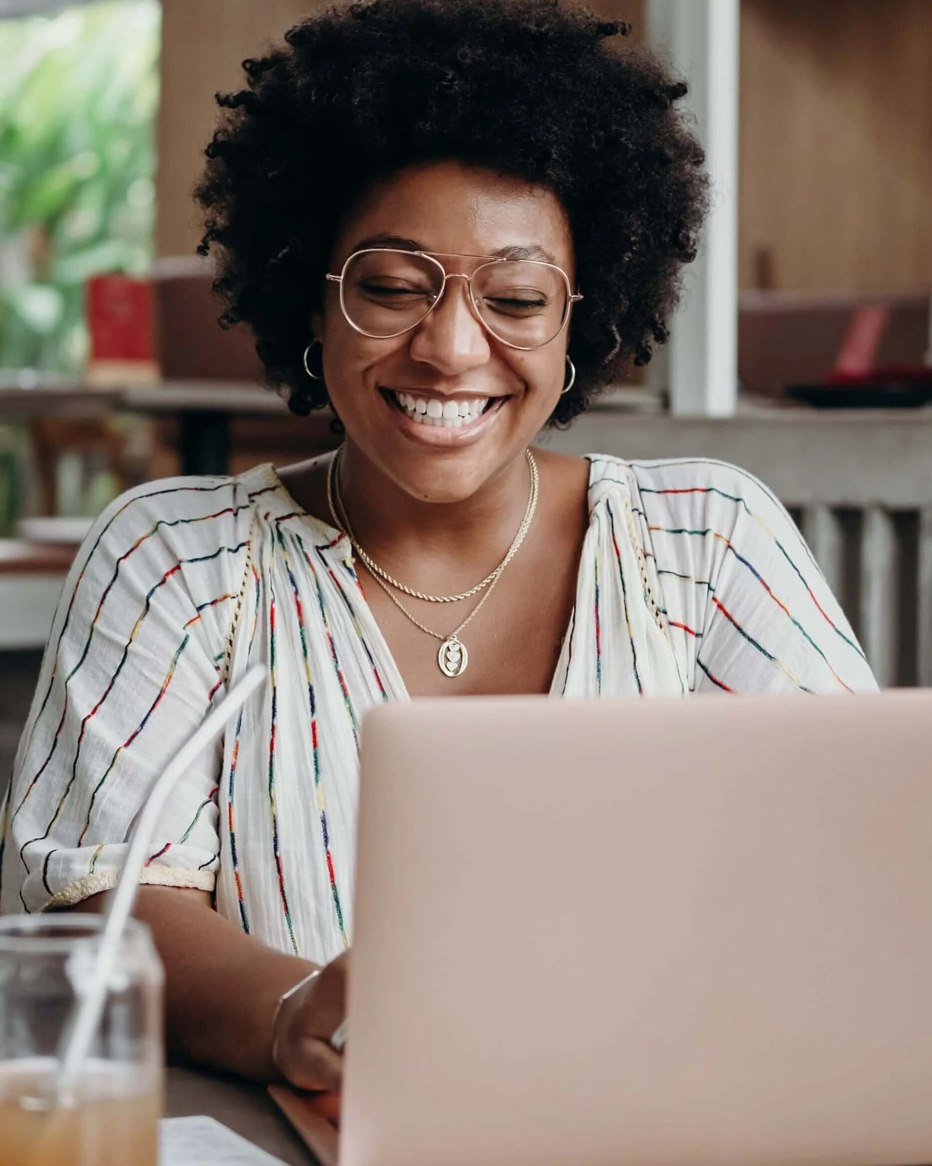 A smiling young lady, sitting at a table with a laptop and a closed red notebook in front of him.