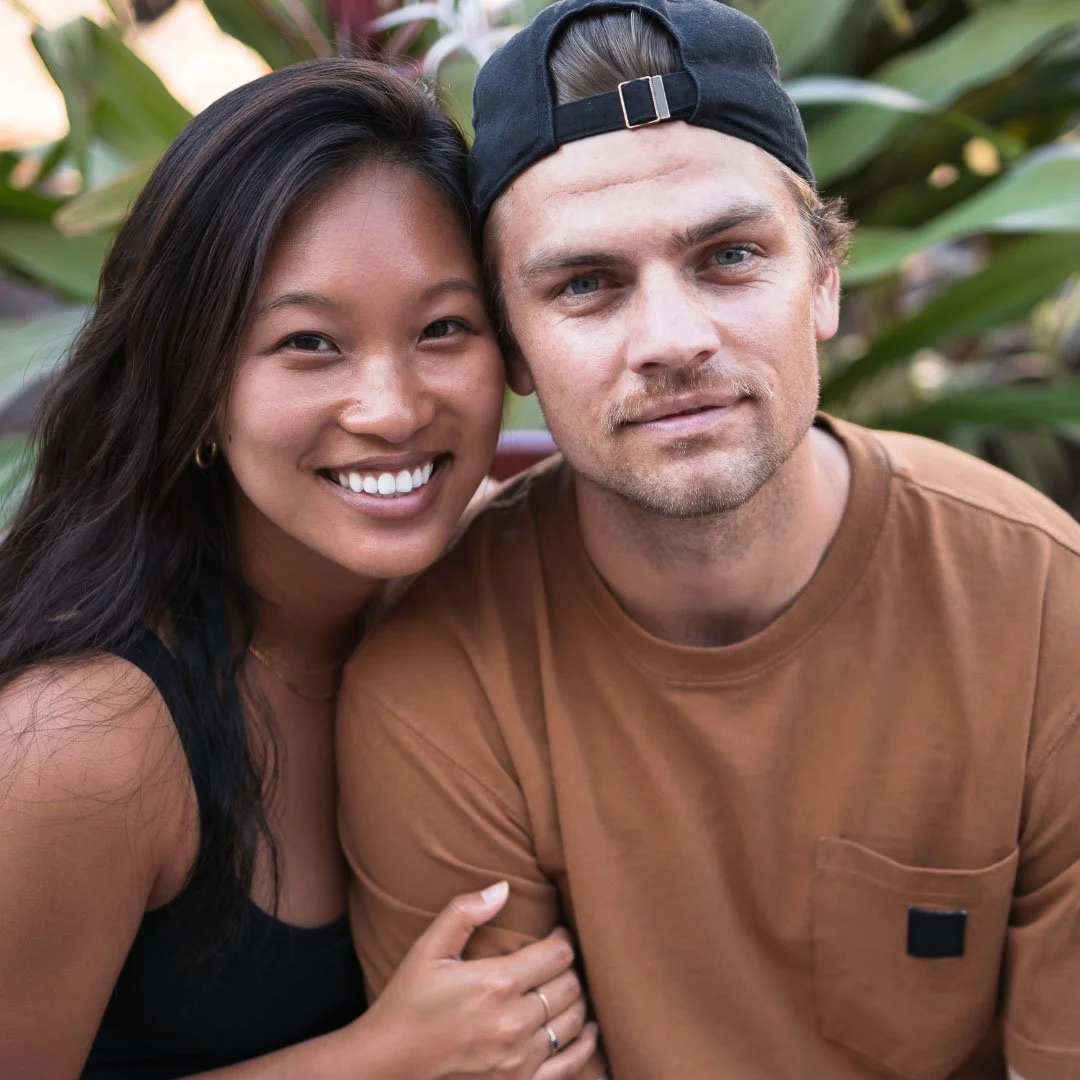 Close-up of a smiling woman with long dark hair and a man with a backwards black cap, sitting outdoors surrounded by greenery.