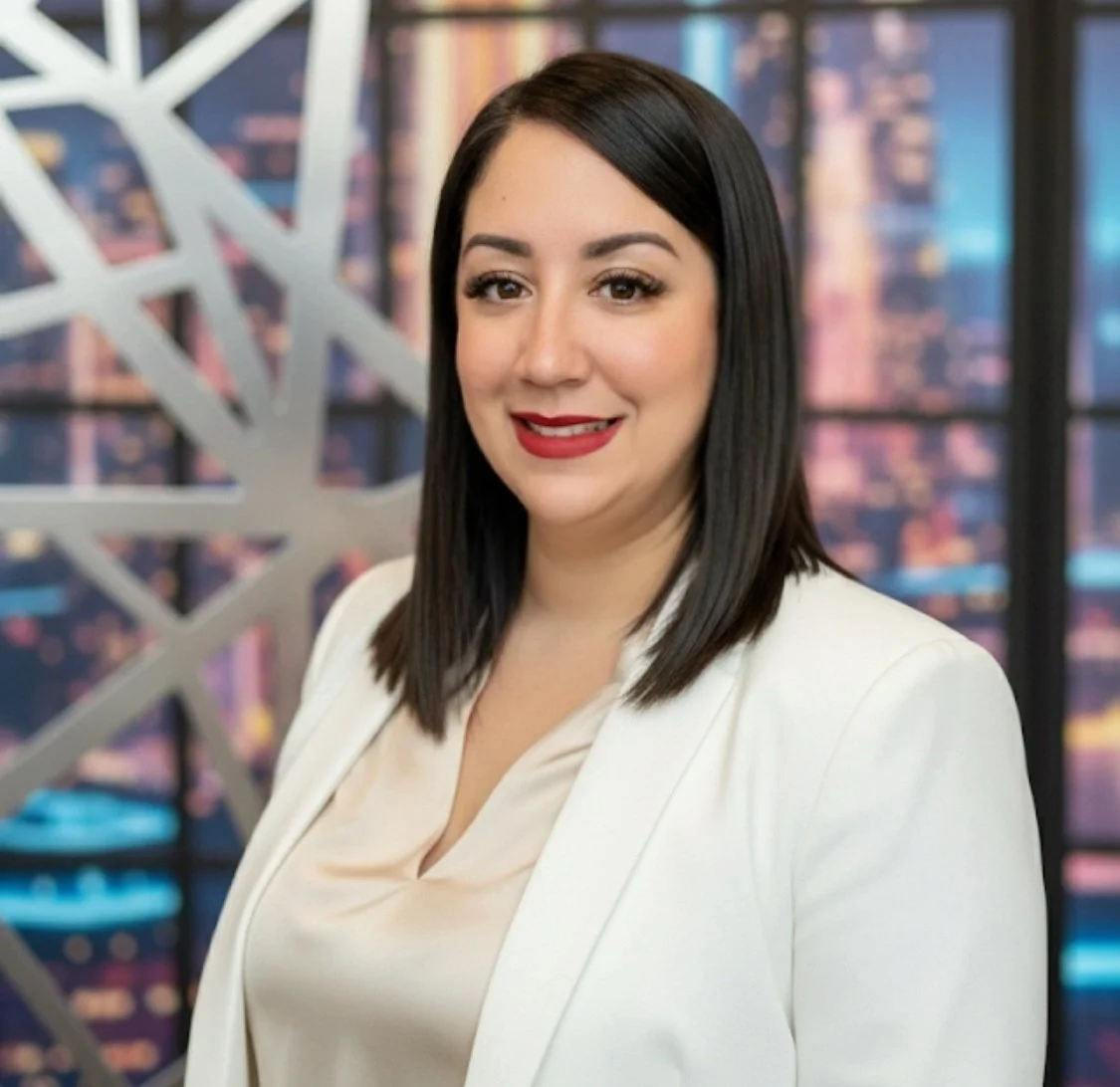 Retrato de una mujer con cabello negro y blusa beige, vestida con saco blanco, en un entorno de oficina con ventana trasera que muestra edificios en la ciudad.