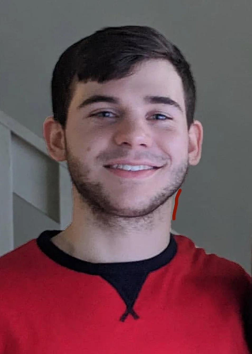 A young man with dark hair, blue eyes, and light skin smiling at the camera, wearing a red and black shirt, standing indoors.