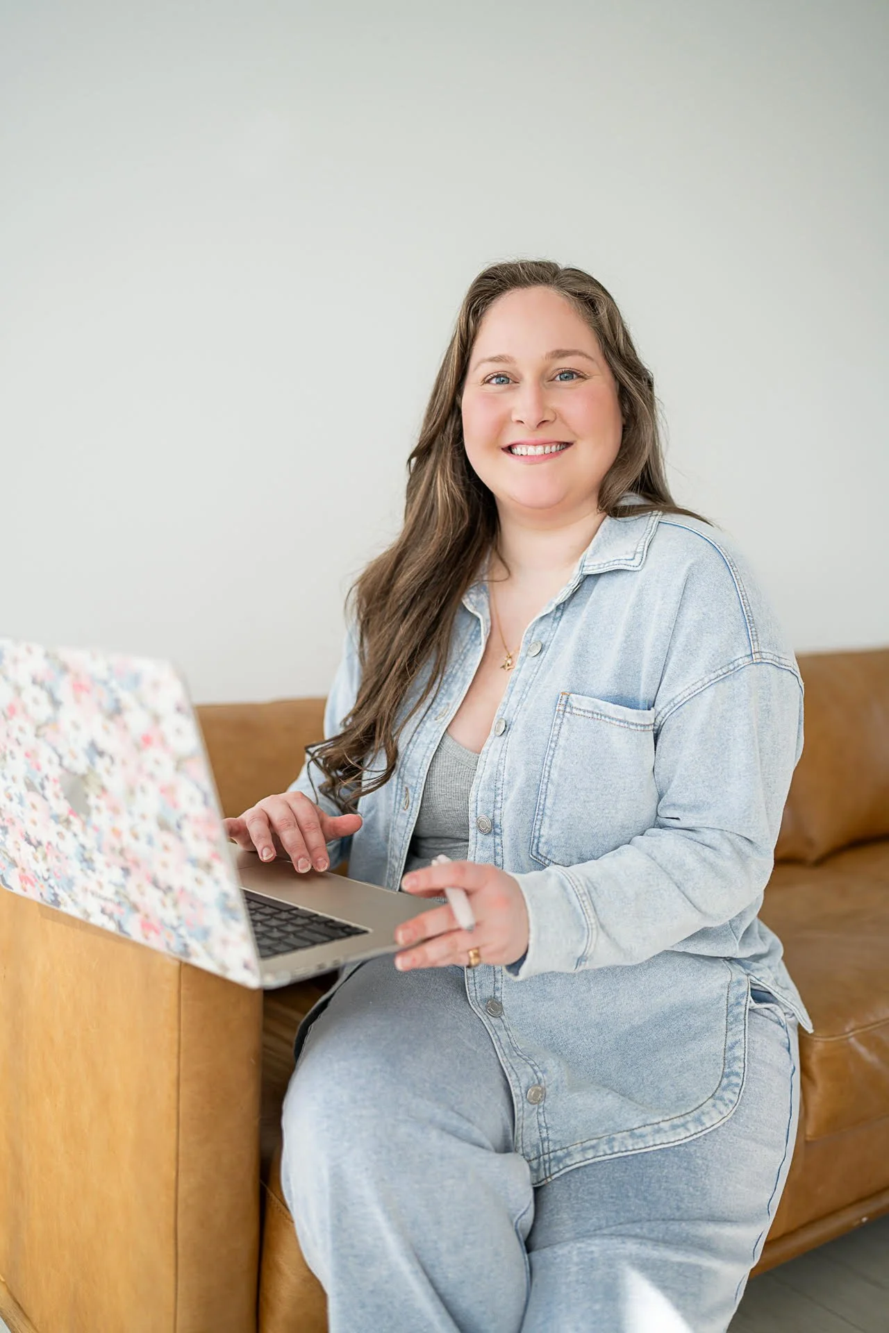 A woman with long brown hair, smiling, sitting on a tan leather couch, using a laptop with a floral cover, wearing a light denim jacket and gray top.