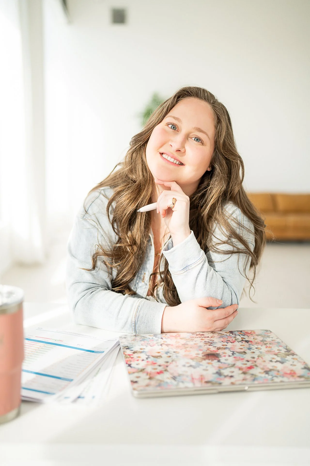 A woman with long wavy brown hair smiling and resting her chin on her hand, sitting at a table with documents and a floral notebook.