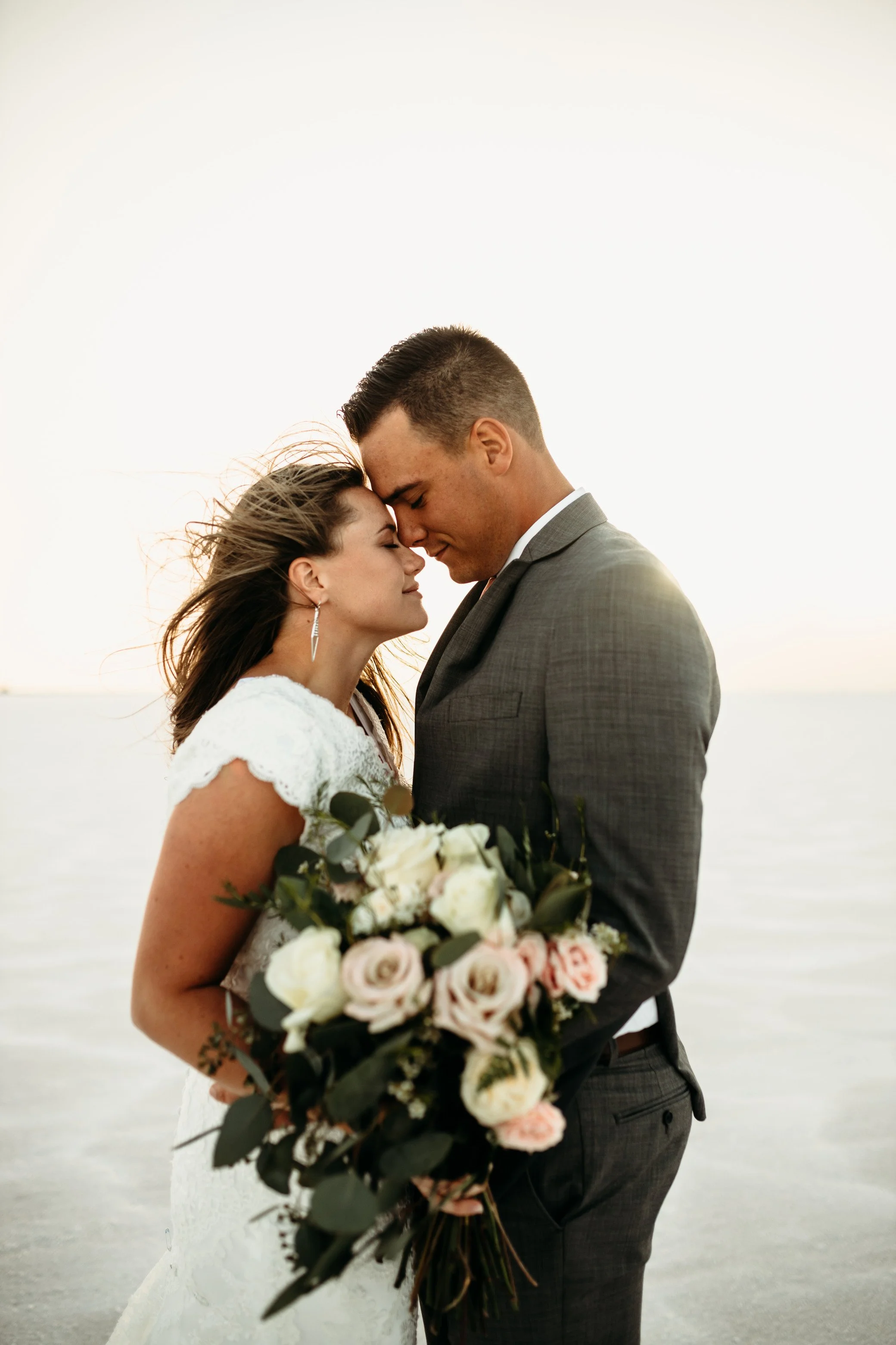A bride and groom with foreheads touching, holding a bouquet of white and pink roses at Salt Flats in Utah