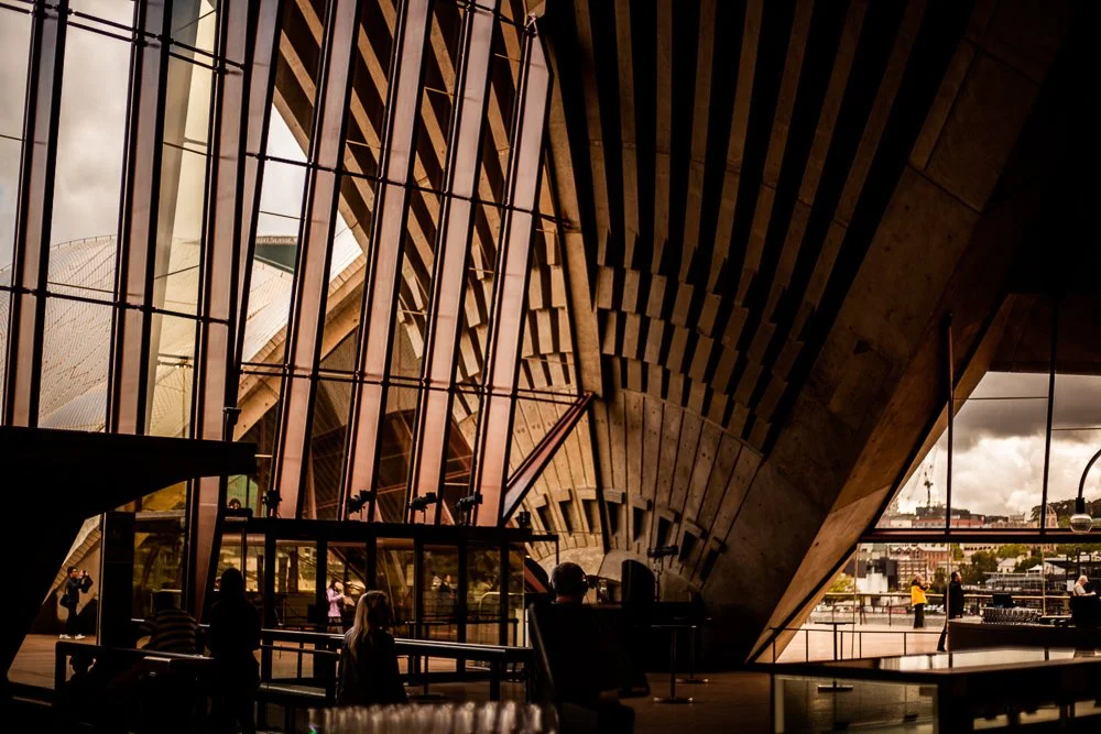 Inside the Sydney Opera House with large glass windows and wooden architectural elements, with people walking and sitting.