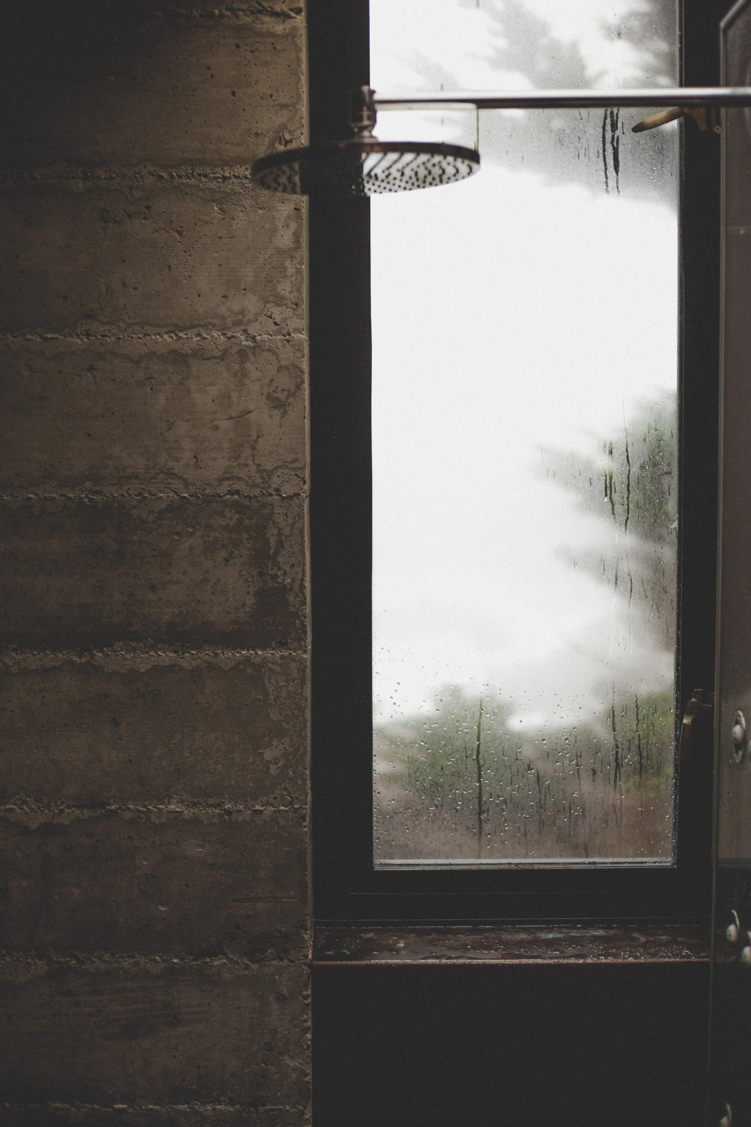A showerhead mounted on a wall next to a rain-streaked window with an overcast sky visible outside.