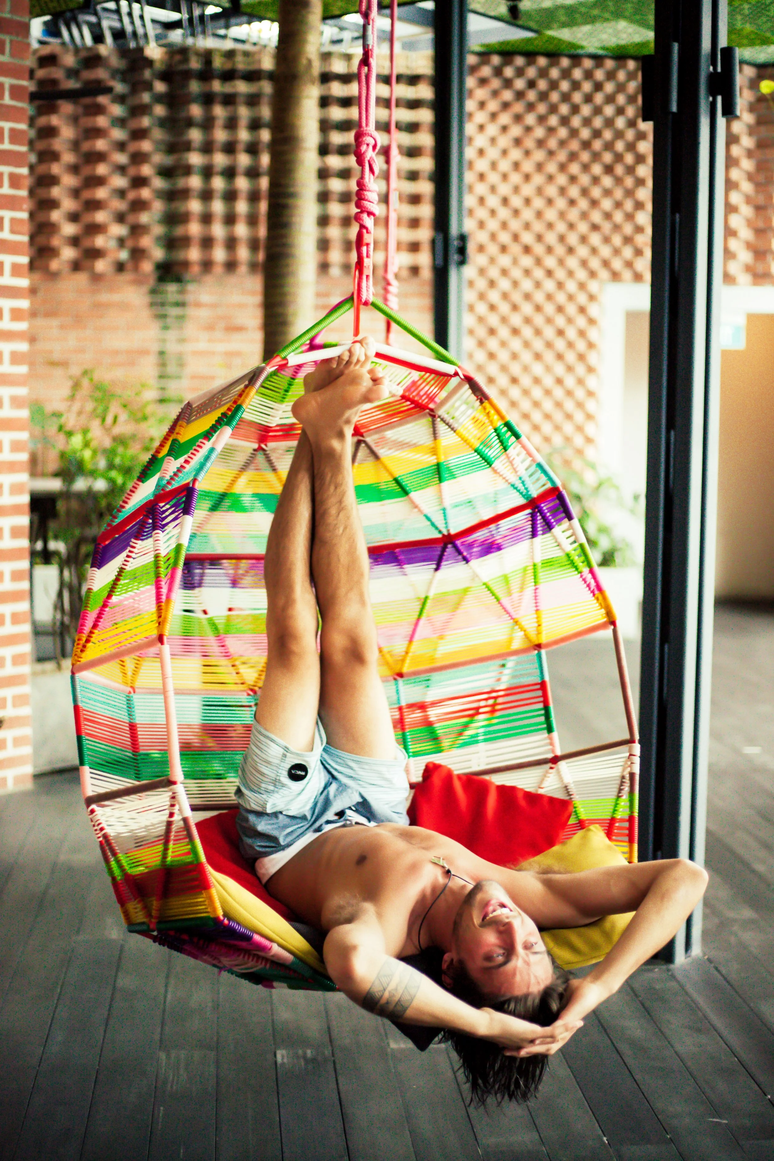 A man lying on a colorful hanging chair with a rainbow pattern, laughing with her head tilted back, in an outdoor or semi-outdoor setting.