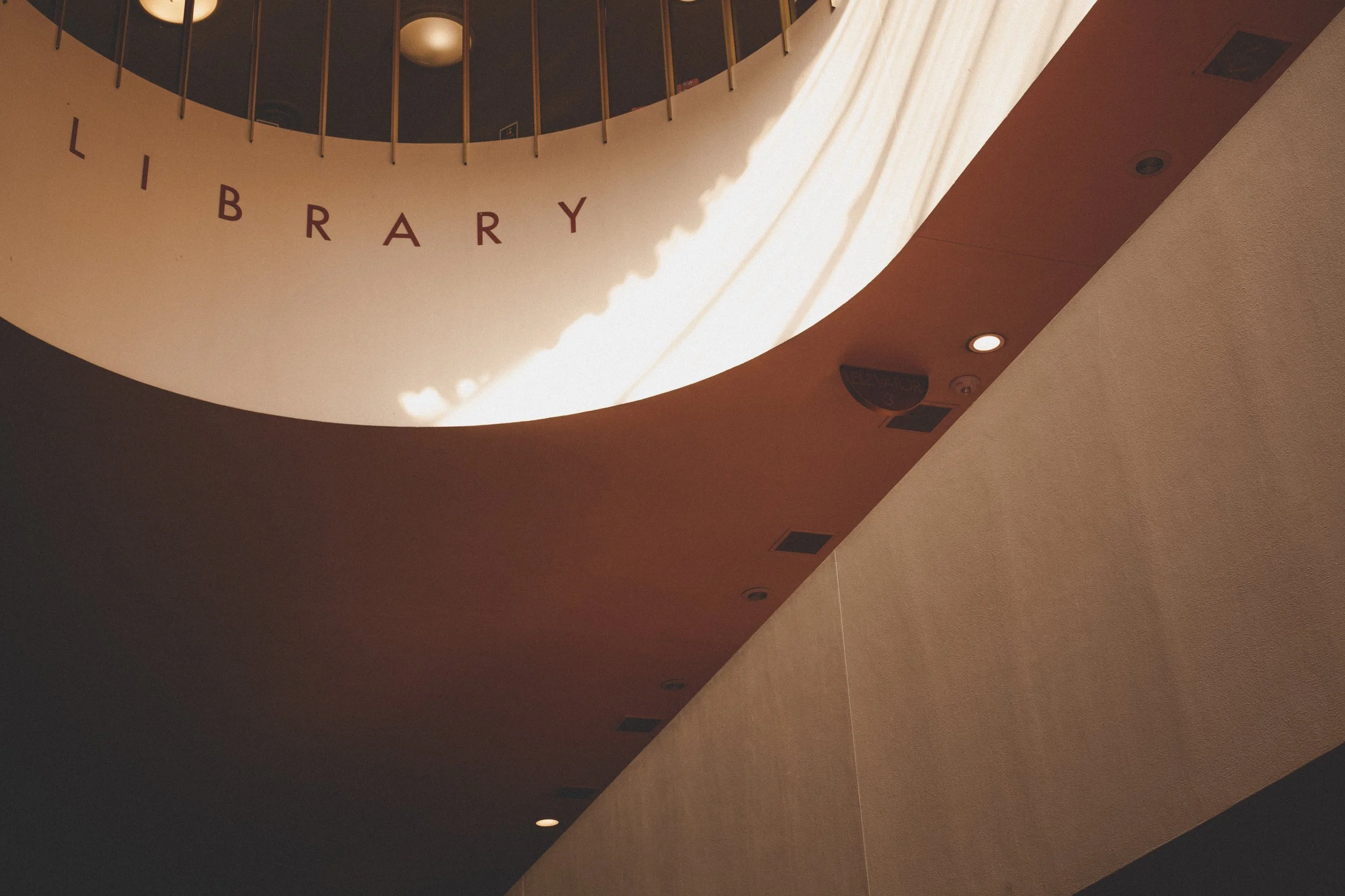 Interior view of a library with a circular opening in the ceiling, sunlight streaming through, and the word 'LIBRARY' spelled out in hanging letters on the balcony above.