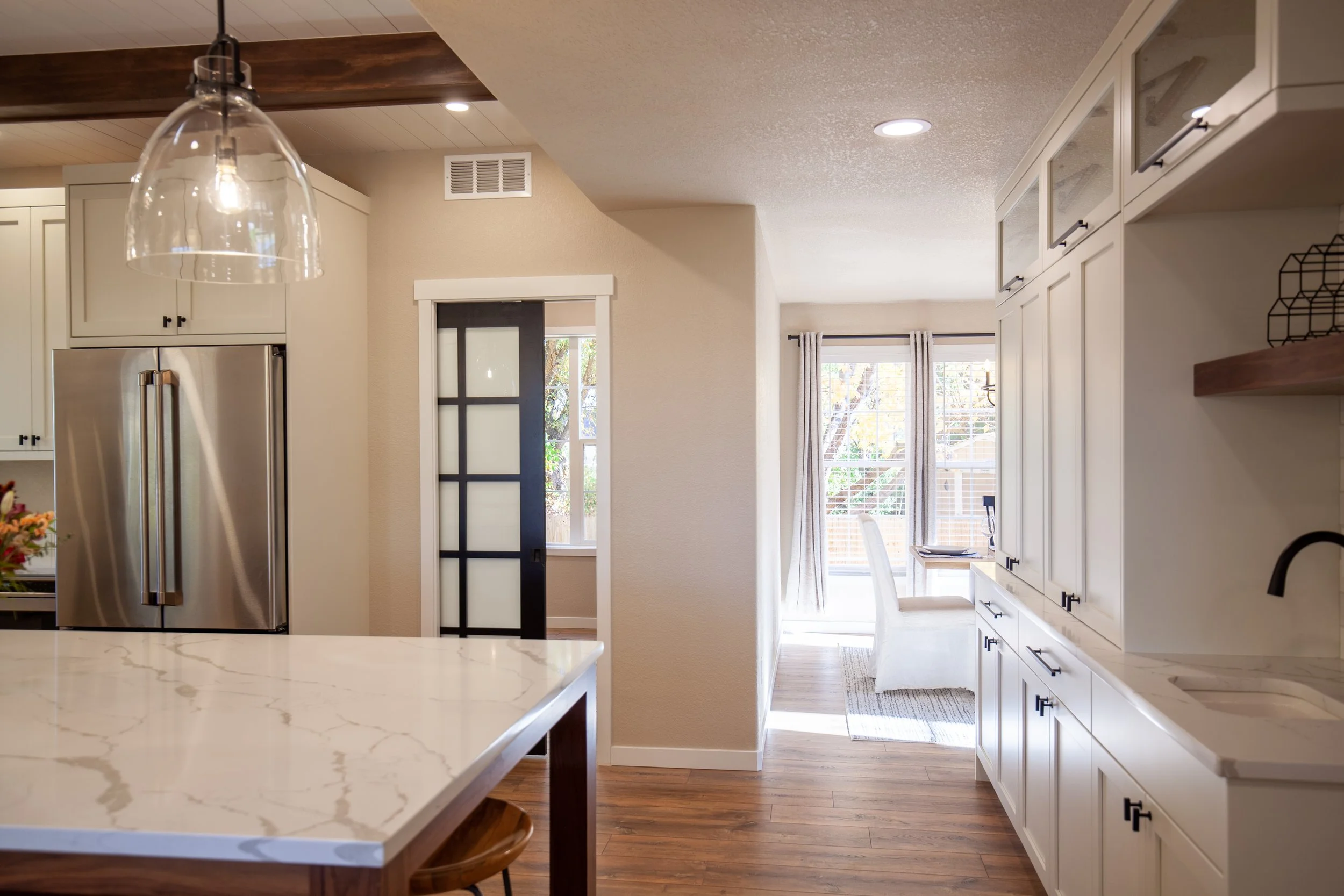 Modern kitchen with stainless steel fridge, marble countertop, pendant light, sliding door, and dining area with large windows.