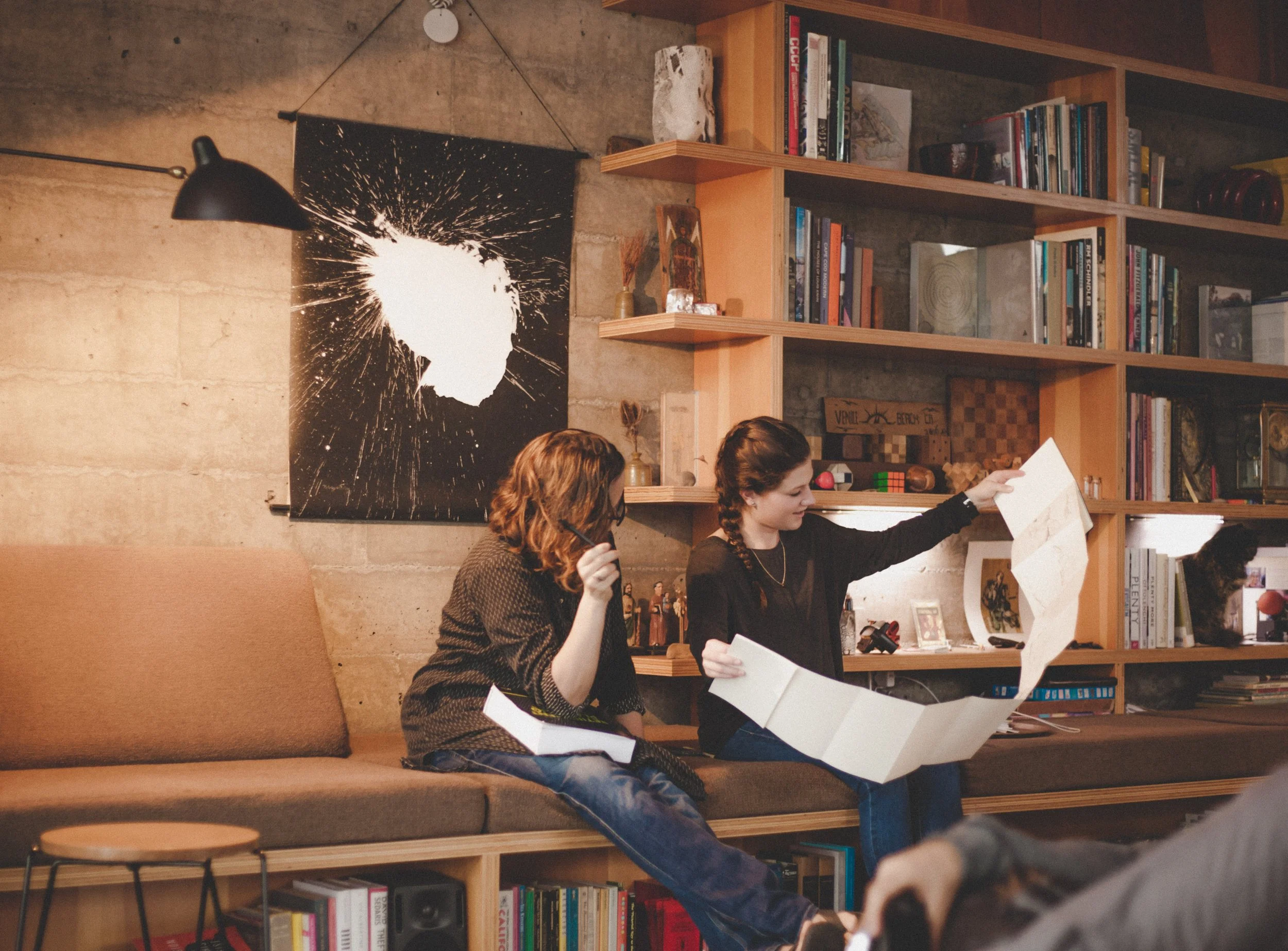 Two young women sitting on a beige couch in a cozy room, one is reading a newspaper while the other is looking at it, with bookshelves filled with books and decorations behind them.