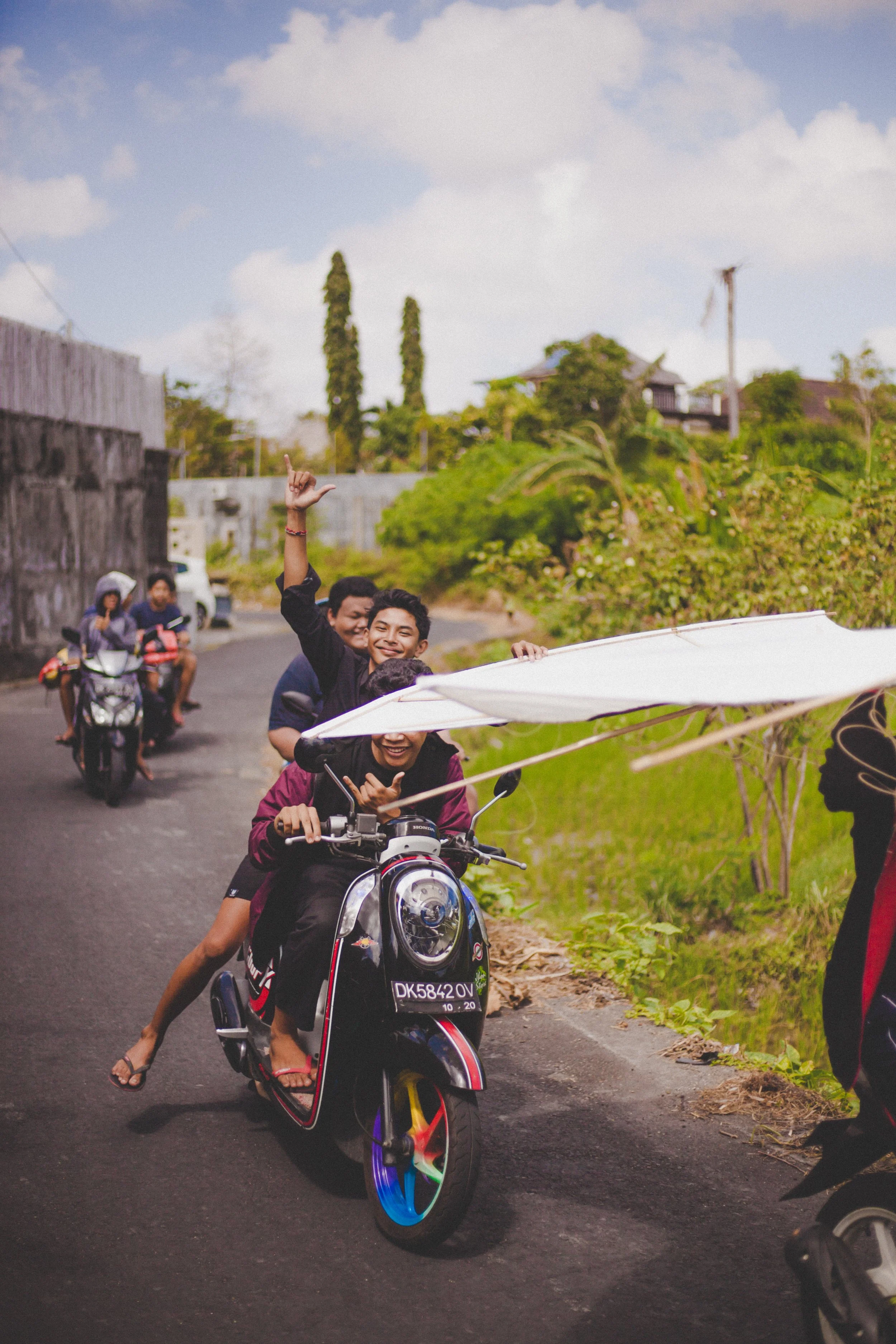 A group of young people riding a scooter along a rural road, with some making playful hand signs and smiling.