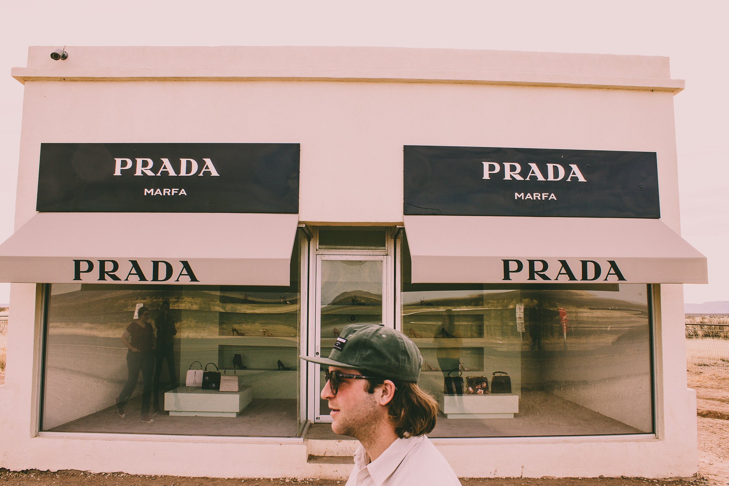 The Prada store in Marfa, Texas with large display windows showing handbags and shoes, with a man in sunglasses and a cap walking in front.