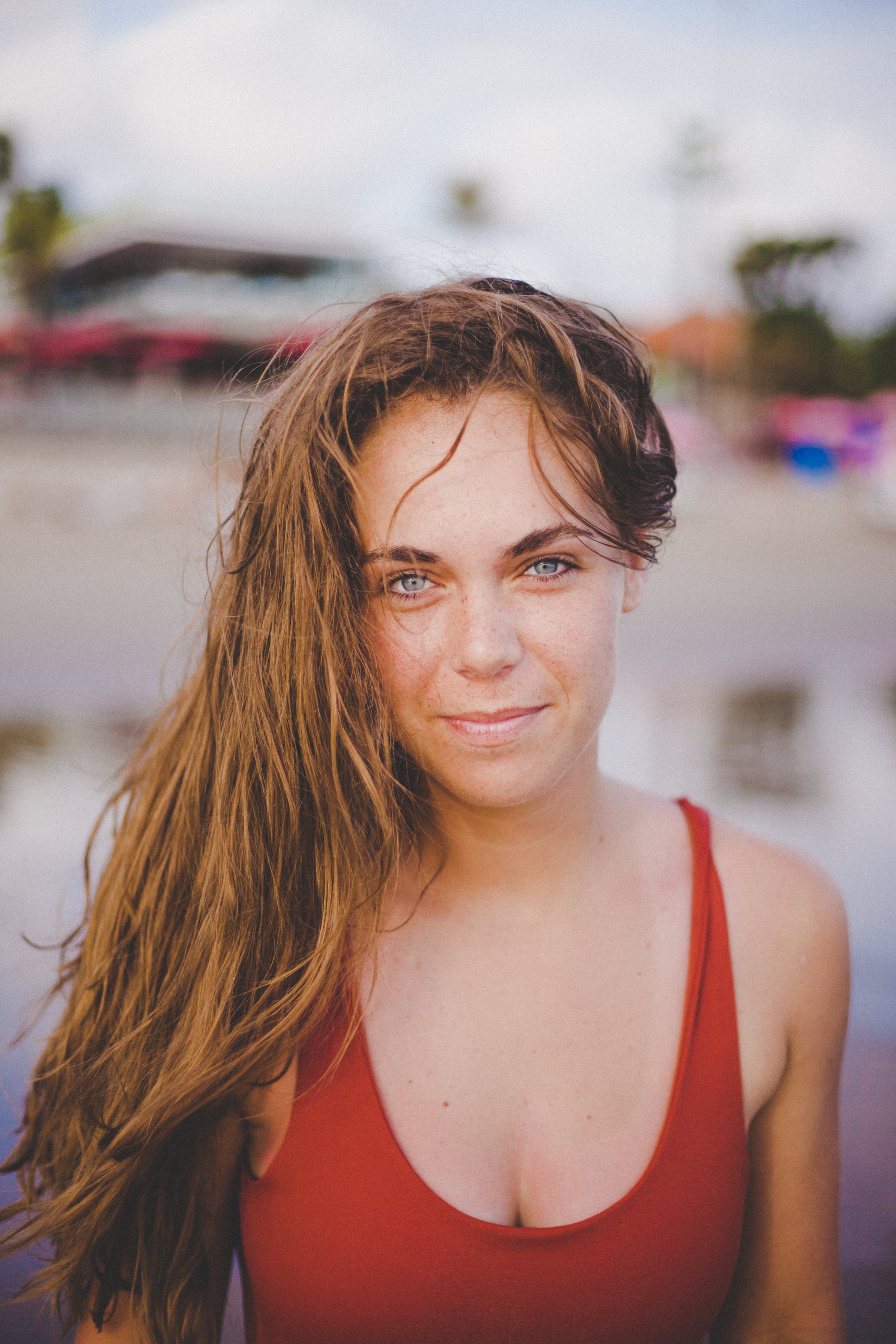 A woman with long, wet, light brown hair and blue eyes standing outdoors near water, wearing a red tank top.