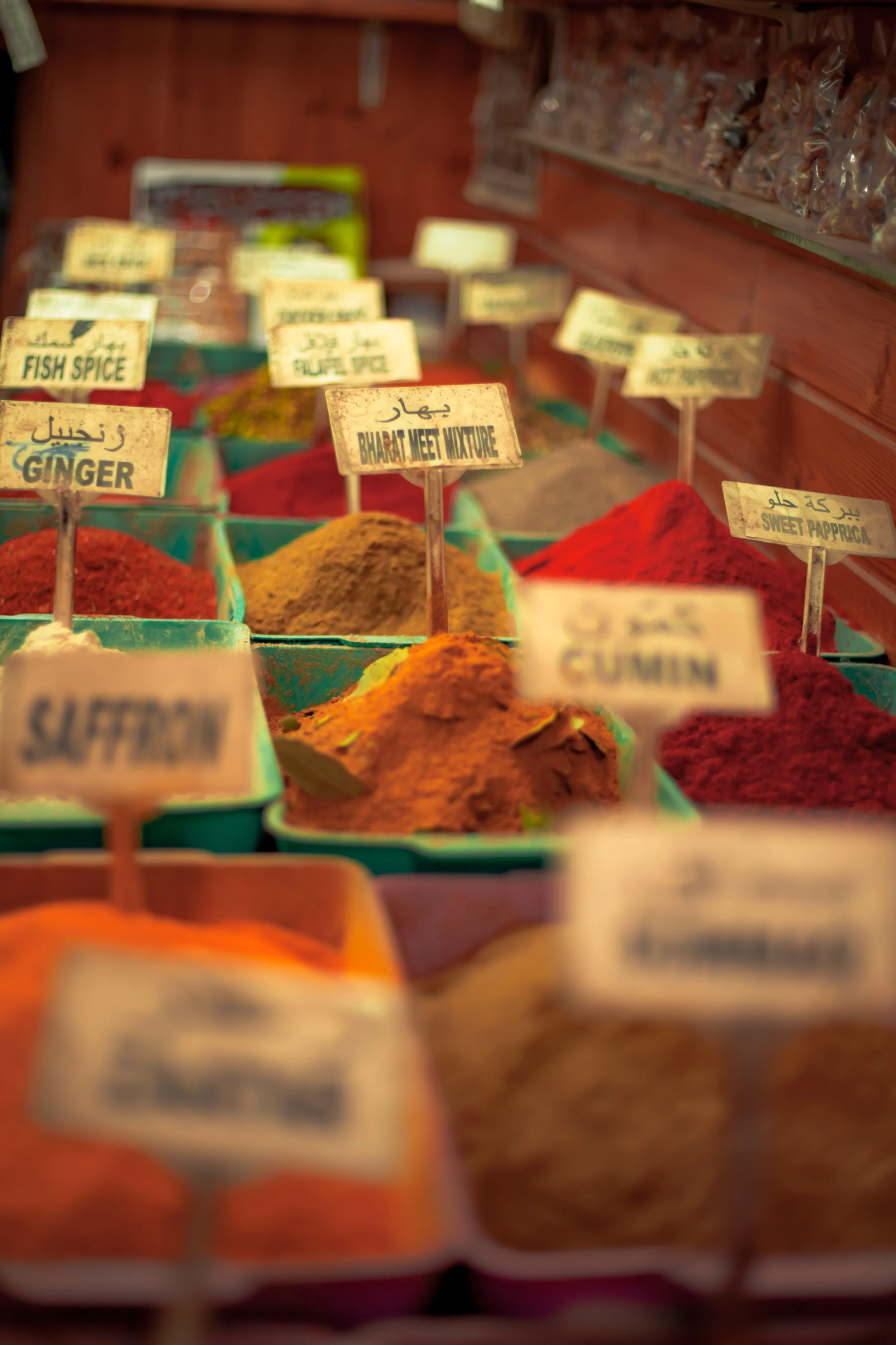 Colorful spices on display in a market stall with labels, including ginger, cumin, sweet paprika, and bharat meet mixture.