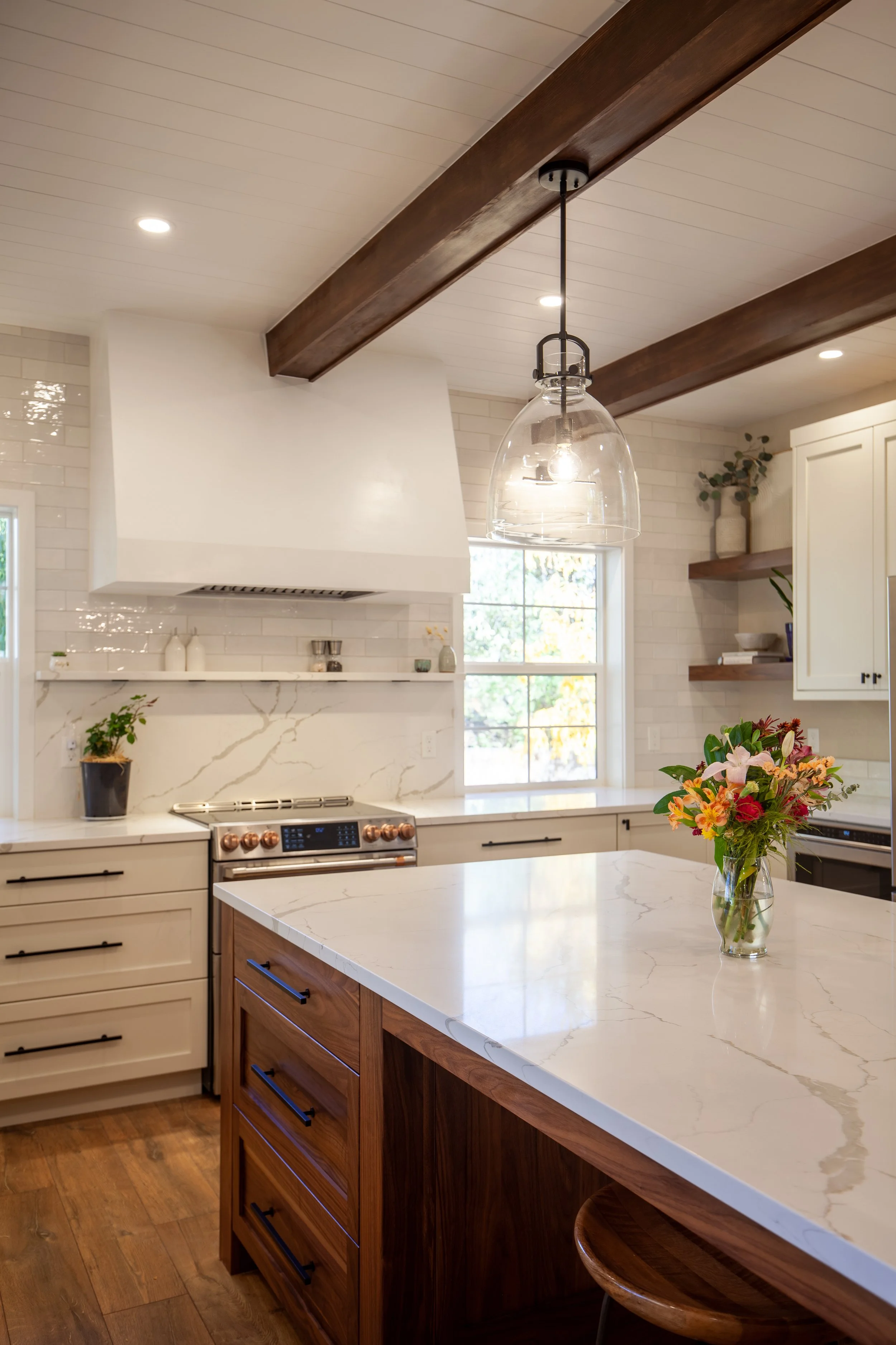 Modern kitchen with wooden beams, white cabinets, and stainless steel appliances. A marble island countertop with a vase of colorful flowers is in the foreground.
