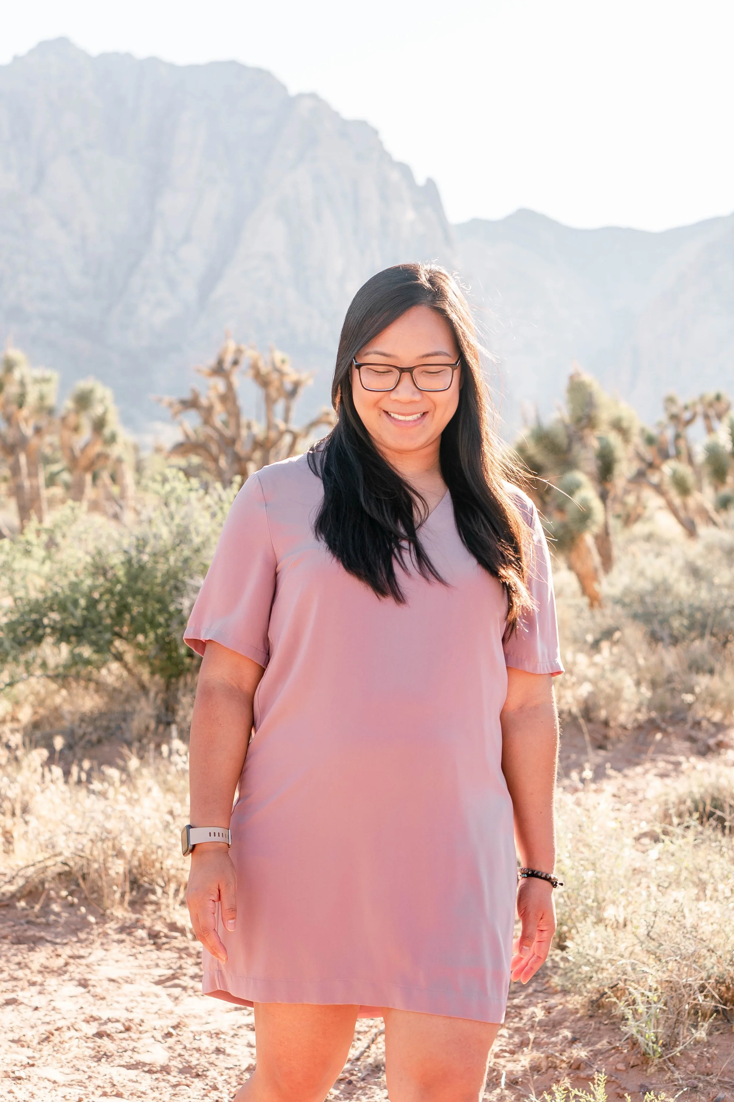 A woman with long dark hair and glasses standing in a desert landscape with mountains and Joshua trees in the background, smiling with her eyes closed.