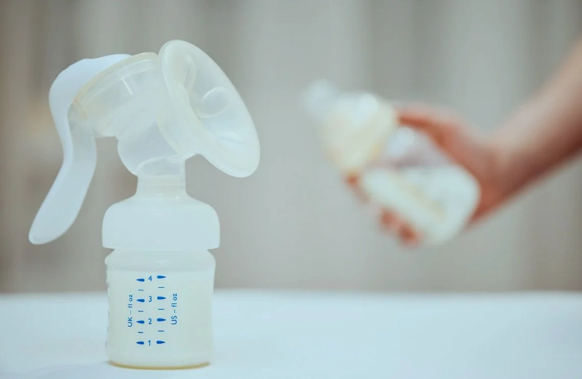 A baby bottle with a breast pump attached, placed on a white surface. A person holding a breast shield with flange in the background.