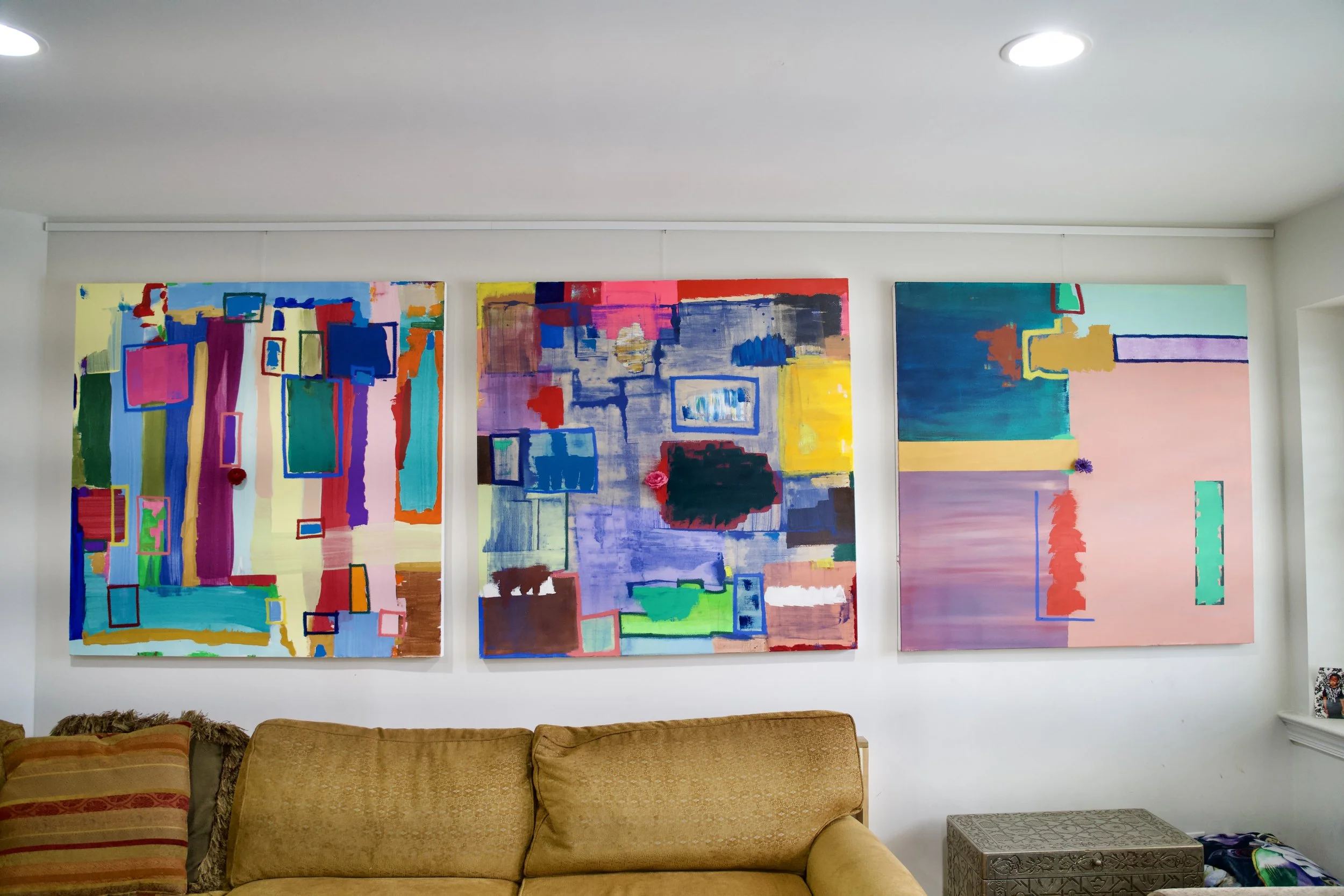 Three large colorful abstract paintings hanging on a white wall in a living room, with a beige couch in front.