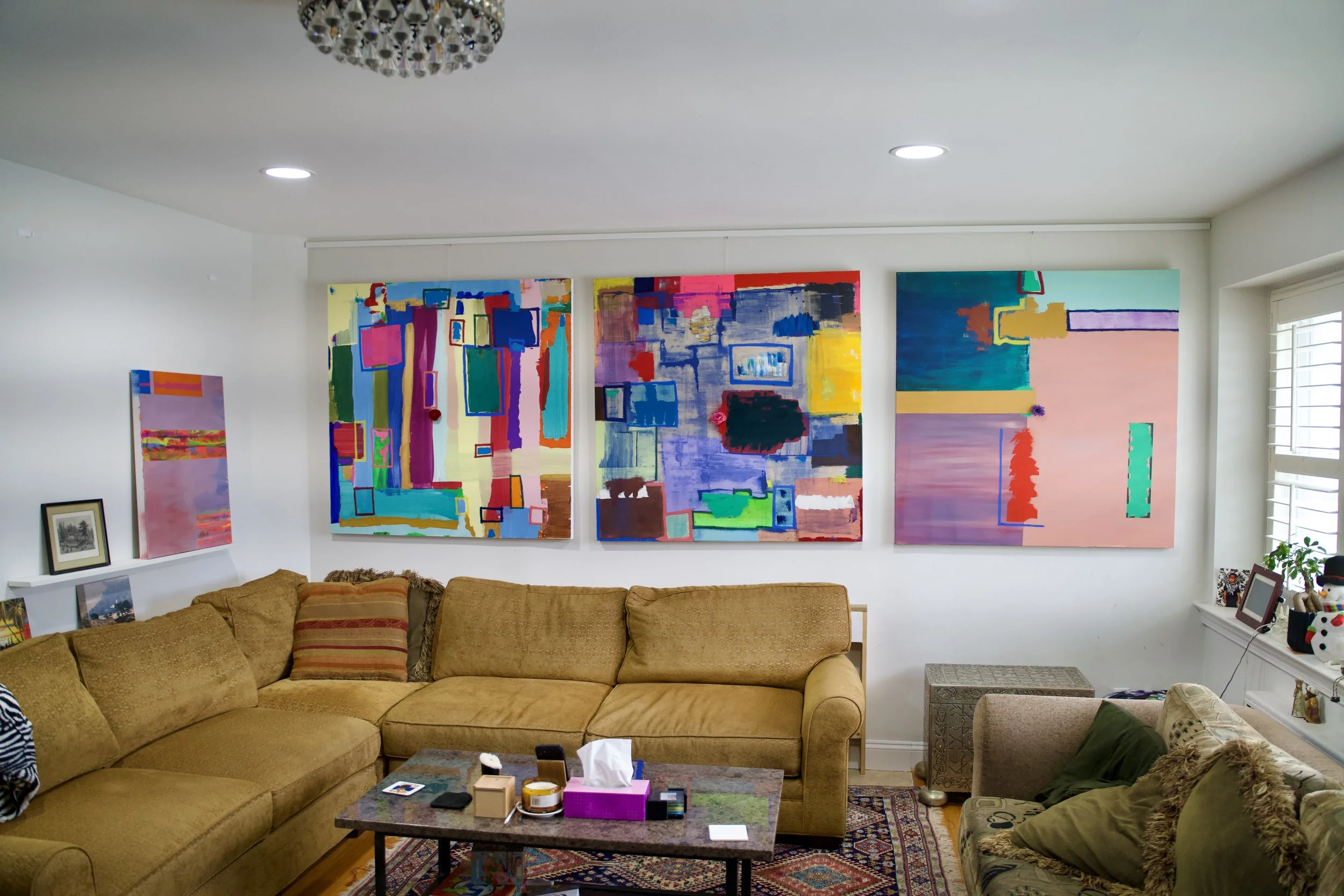 Living room with abstract colorful paintings on the wall, a beige sectional sofa with cushions, a coffee table with items on it, and a window with blinds.