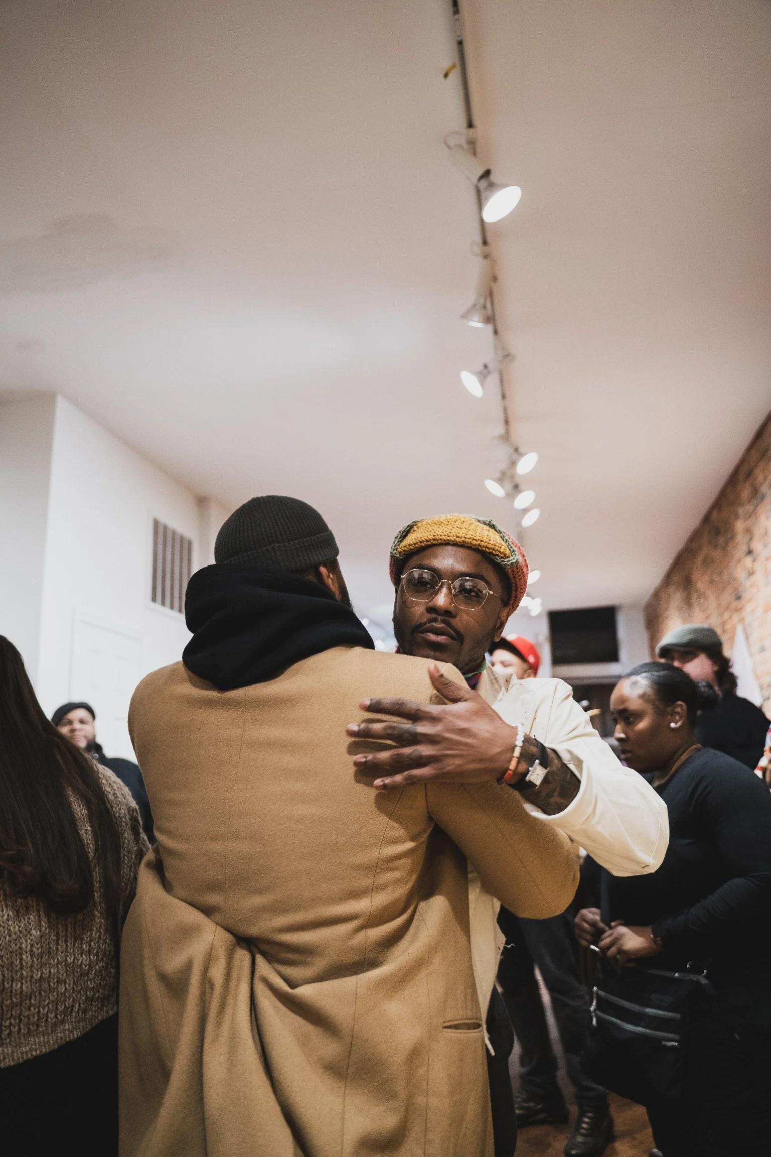 Two men hugging at a social gathering, with several other people in the background in a room with a brick wall on the right and track lighting on the ceiling.