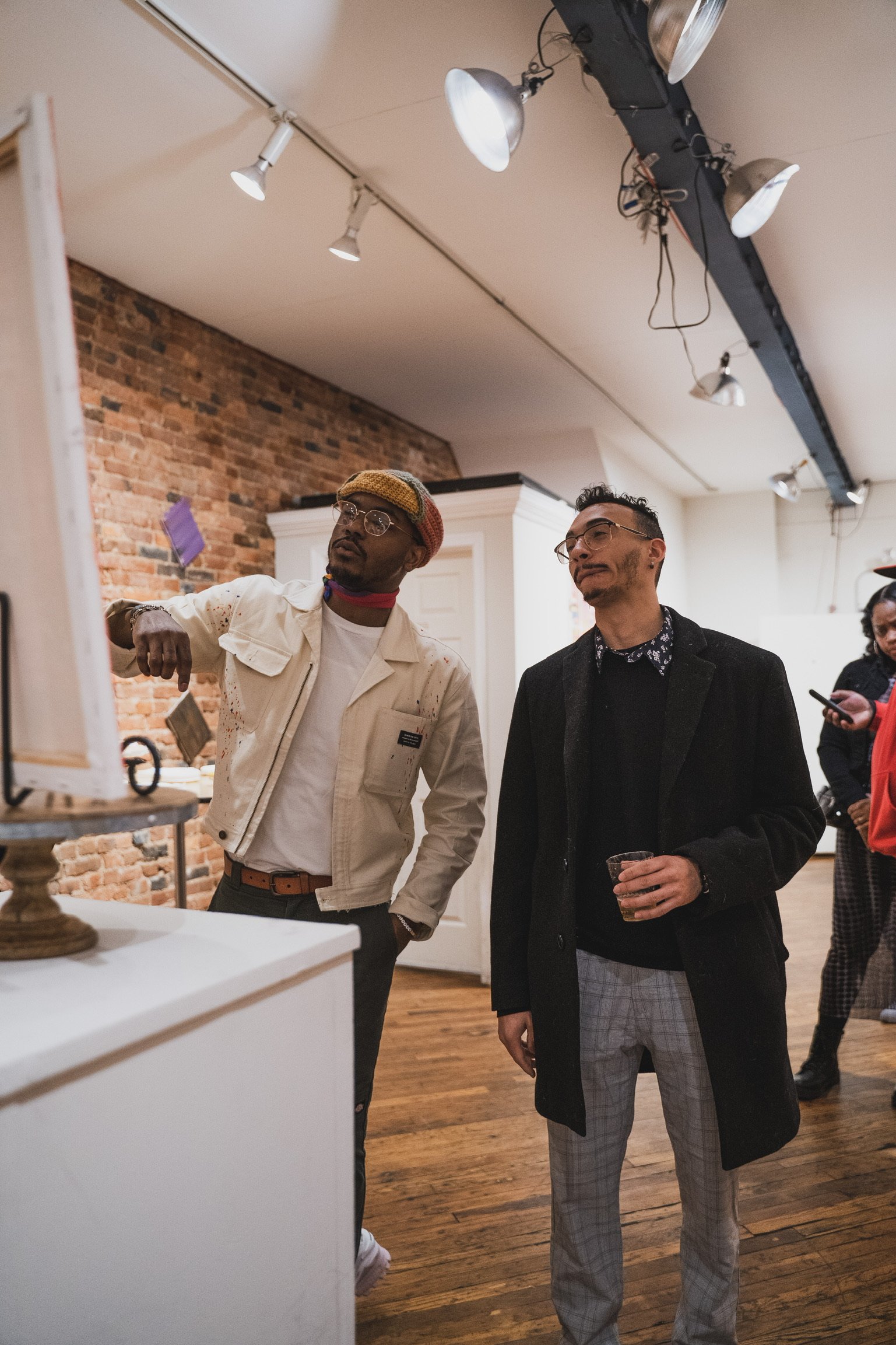 Two men are standing and looking at an art display or exhibit in a room with a brick wall and wooden flooring. One man is wearing a white jacket, glasses, and a colorful knit hat, while the other is in a black coat and plaid pants, holding a drink.