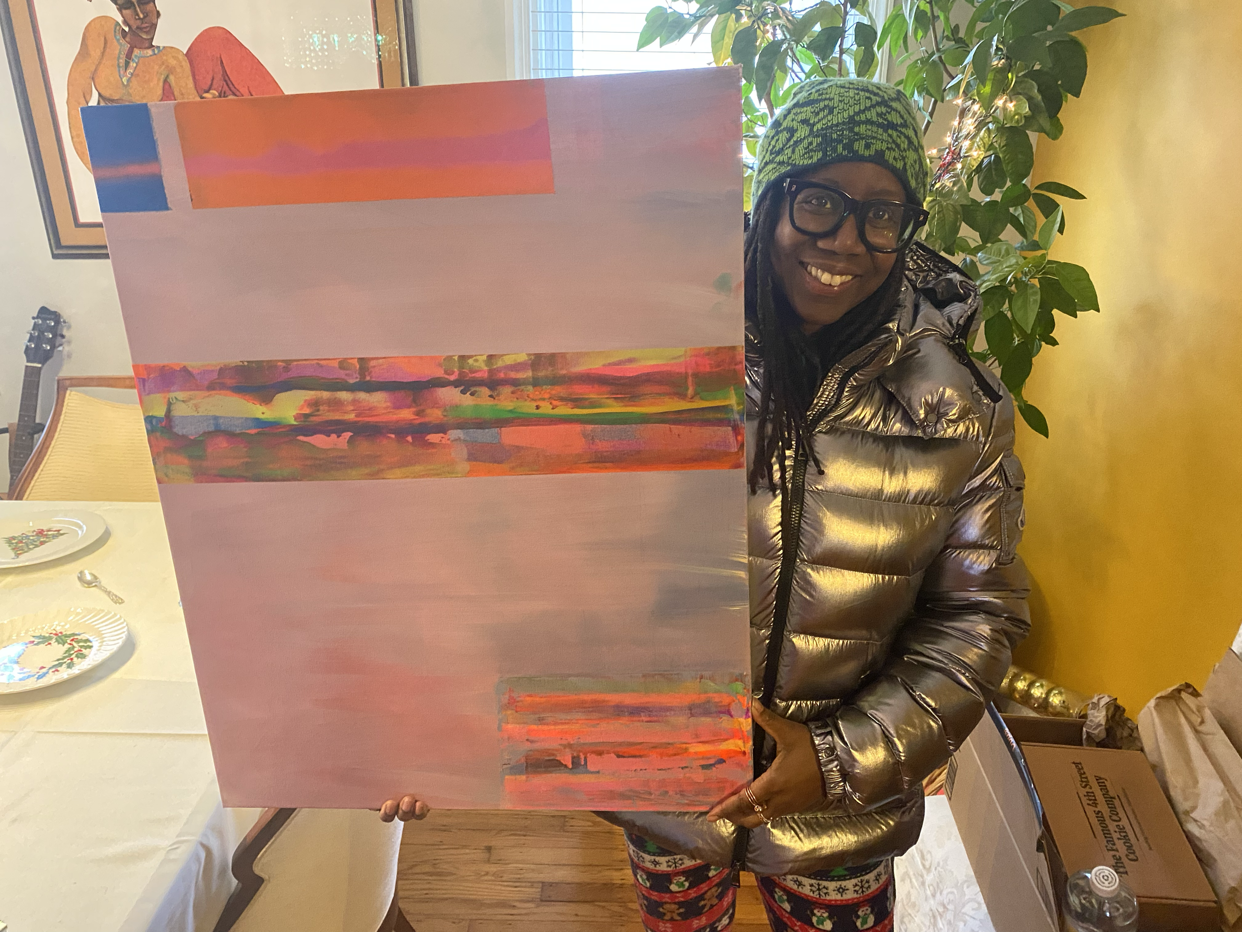A woman wearing glasses, a green and black beanie, and a shiny silver jacket is smiling while holding a colorful abstract painting. She is standing in a room with a yellow wall, a large green plant, and holiday plates on a table.