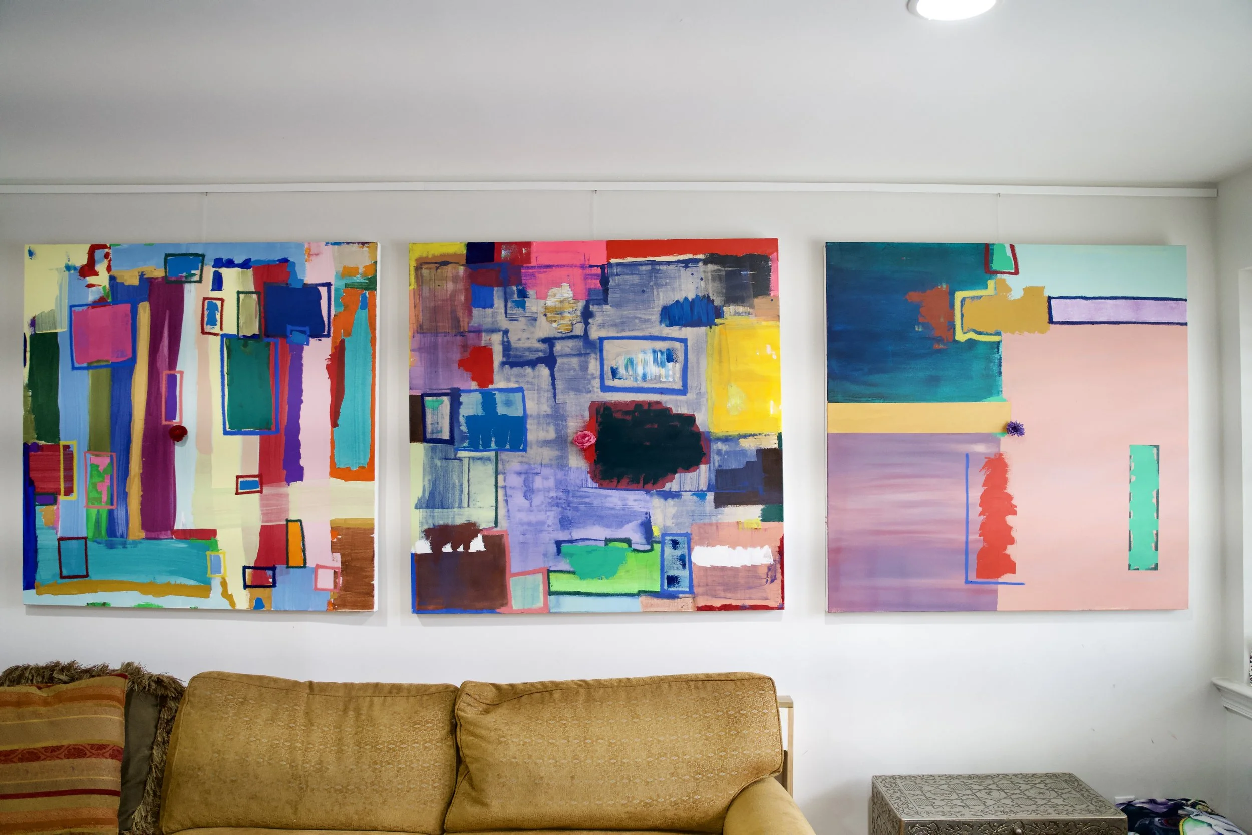 Three abstract paintings hanging on a white wall above a brown couch in a living room.