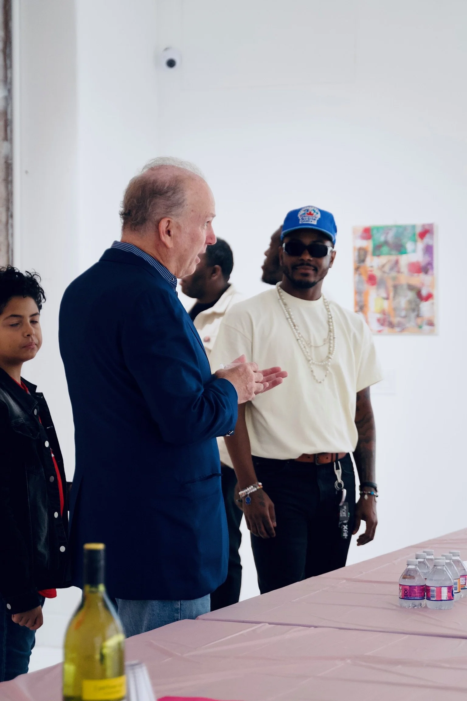 A group of people standing around a table with bottled water and a yellow bottle, in a white gallery space with artwork on the wall.
