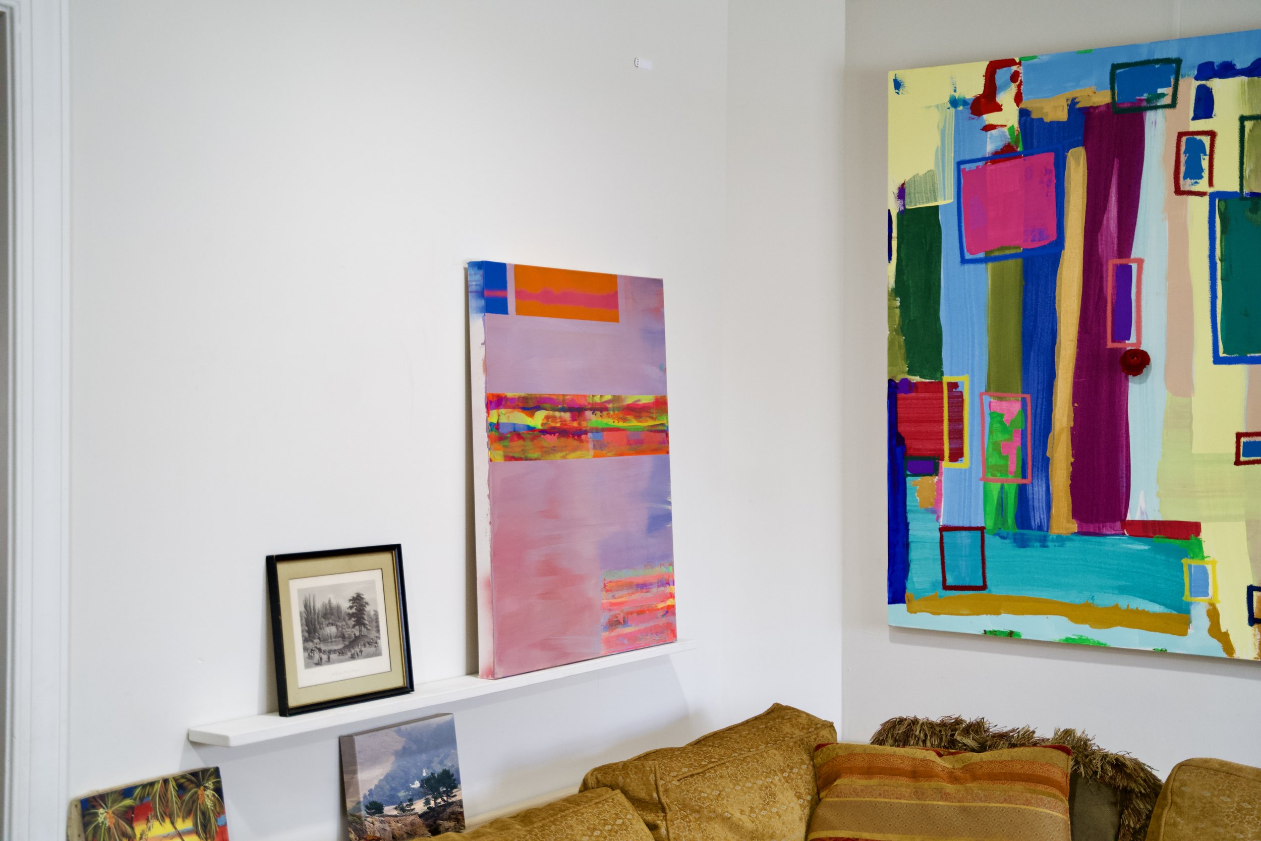 Interior of an art gallery with colorful paintings on the white walls, including a large abstract piece with bright colors and various rectangular shapes, a pink and orange abstract painting, framed black and white artwork, and framed landscape photo