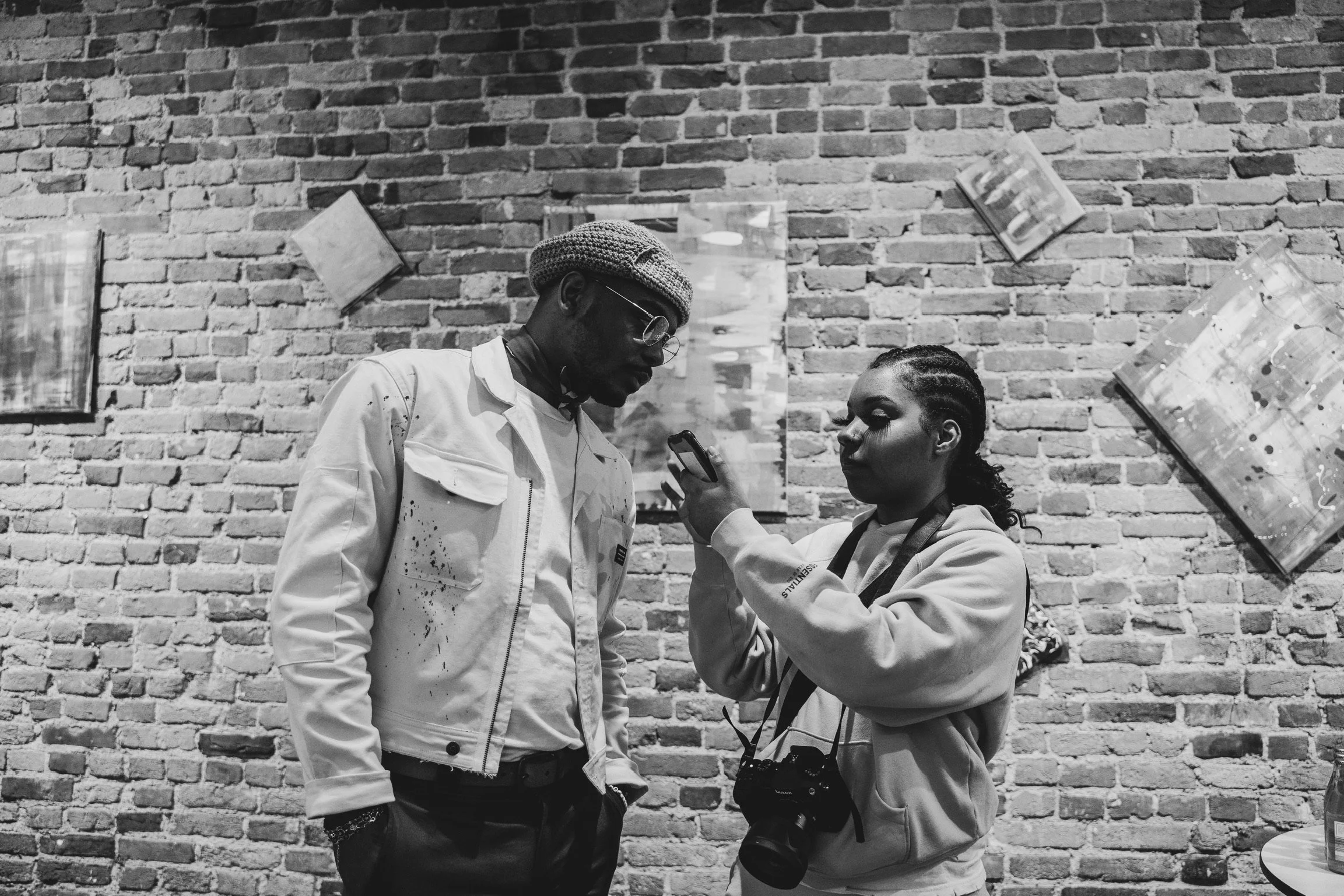 A black and white photo of two people standing in front of a brick wall. The person on the left is wearing glasses, a scarf, and a light-colored jacket. The person on the right, who is holding a smartphone, has a camera hanging from their neck, and i