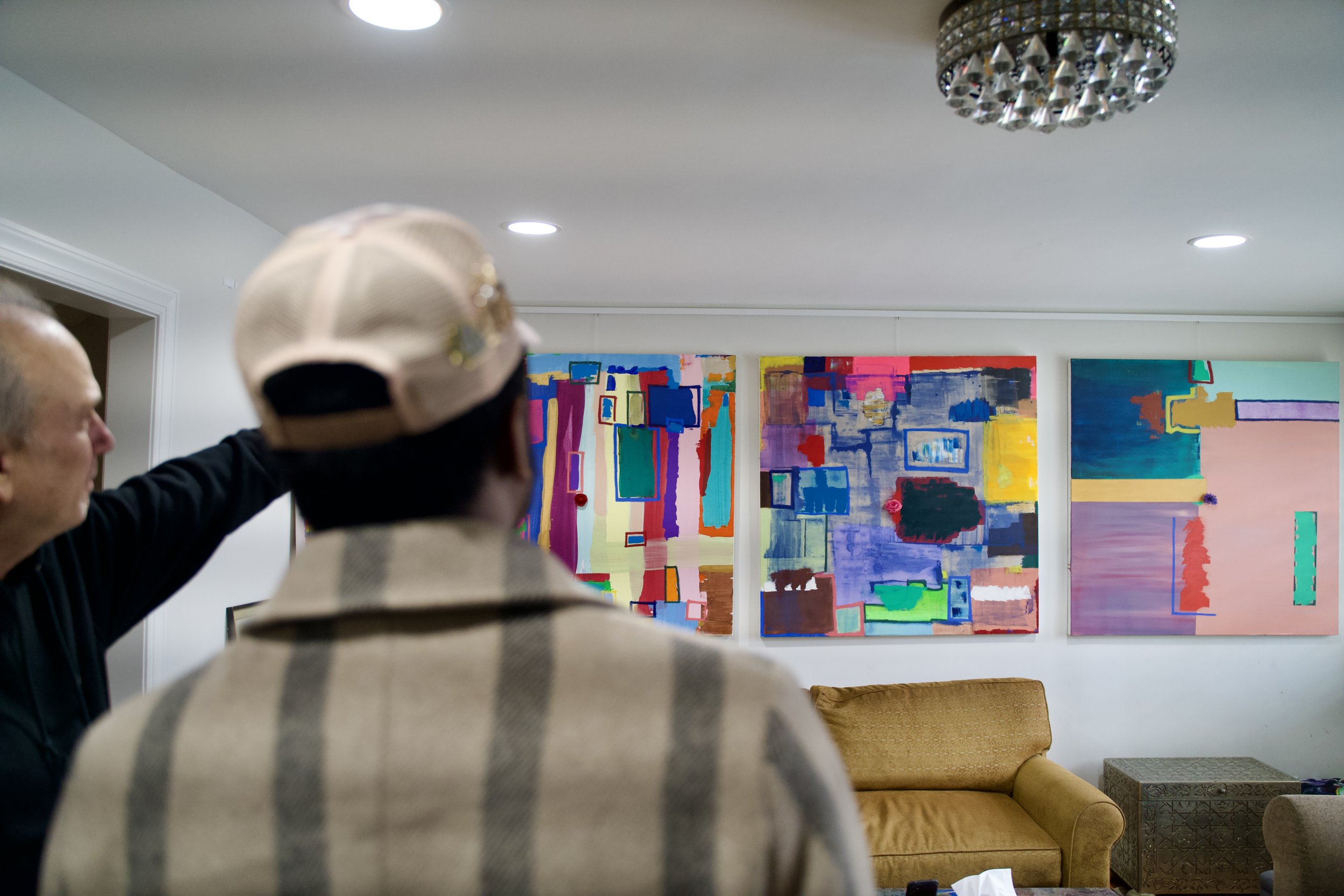 Two men observe colorful abstract paintings hanging on a white wall in an art gallery or living room.