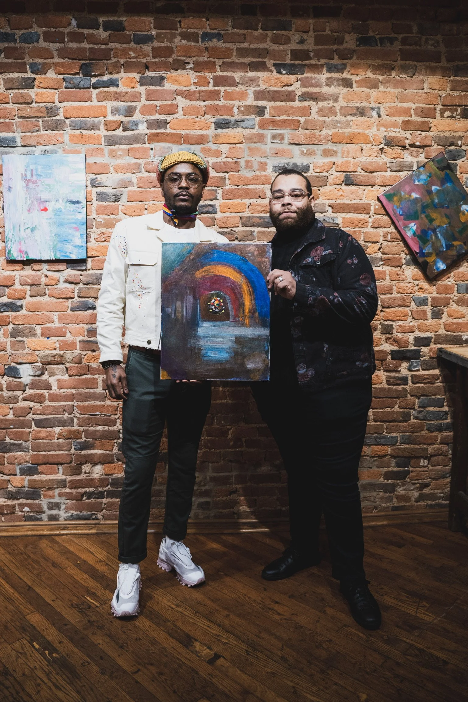 Two men standing in front of a brick wall holding a colorful abstract painting. One man is wearing white sneakers, a white jacket, and glasses. The other man is dressed in black and has a beard. Two additional abstract paintings are hanging on the br