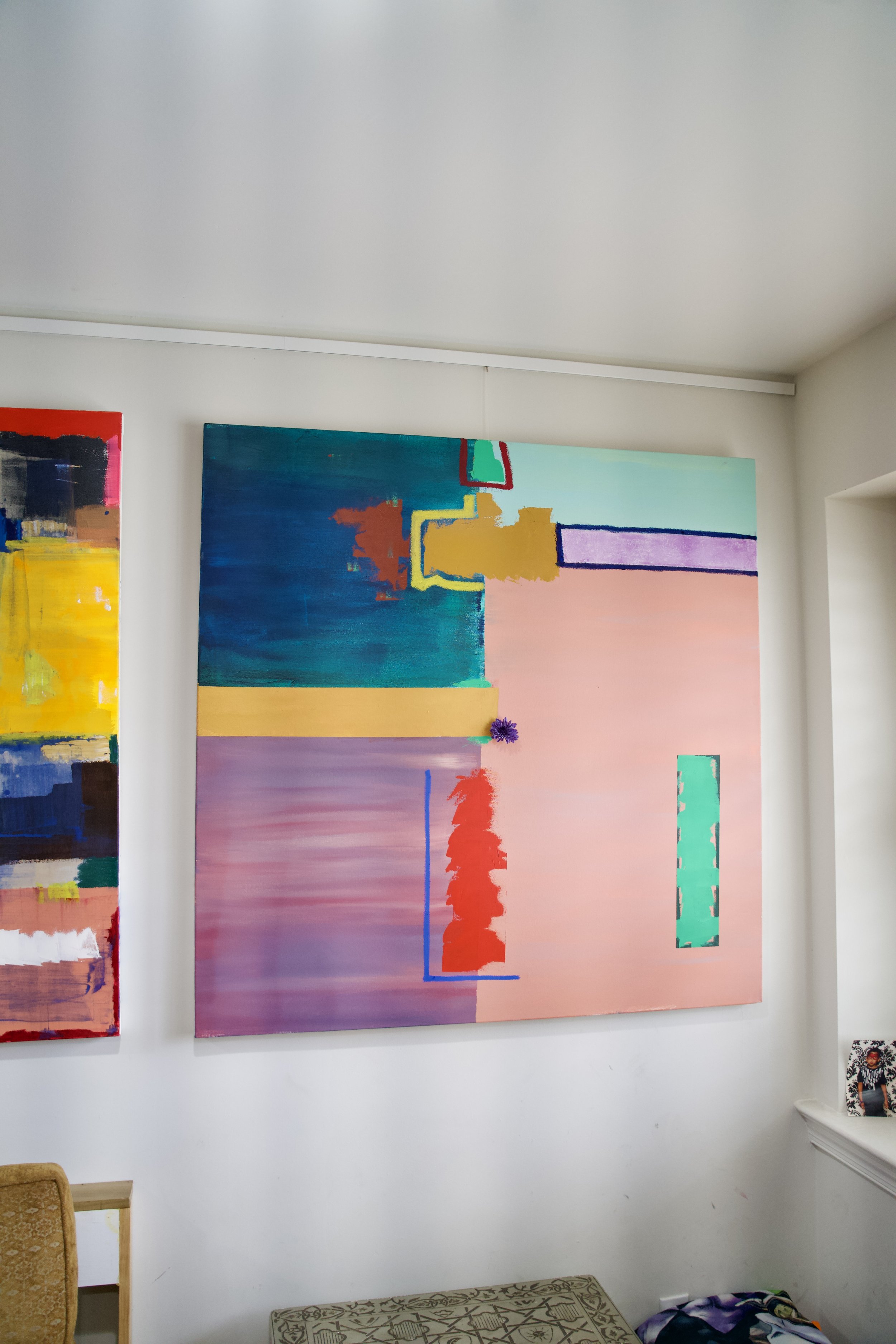 Colorful abstract painting on a gallery wall with a blue, pink, yellow, and teal color palette, featuring geometric shapes and textured brushstrokes.