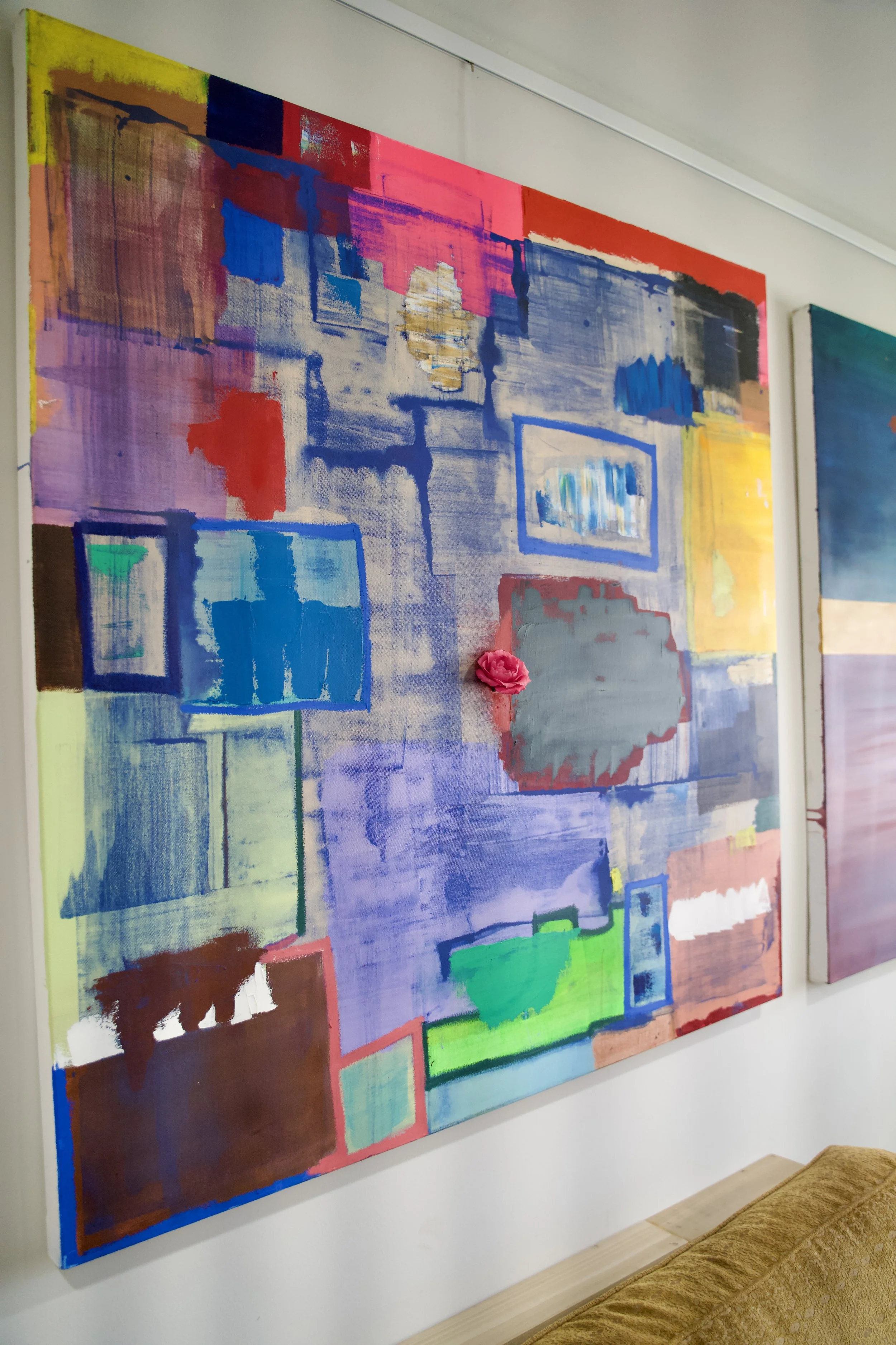 Colorful abstract painting with rectangles and brushstrokes in shades of blue, purple, red, yellow, green, and brown, featuring a small pink flower embellishment, hanging on a white wall.