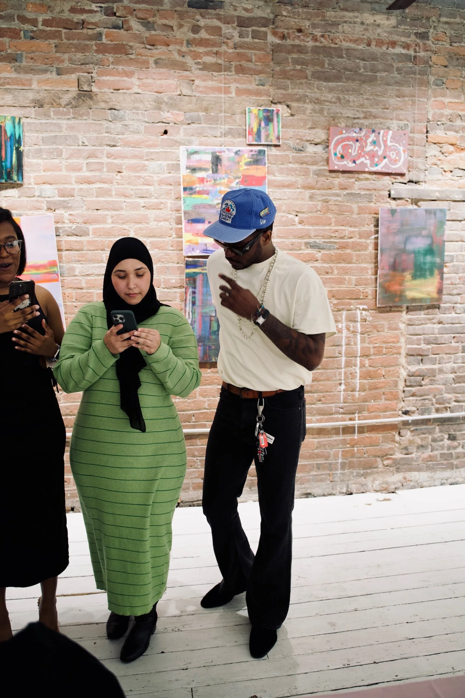 Three people standing in front of a brick wall decorated with colorful abstract paintings, all looking at their phones