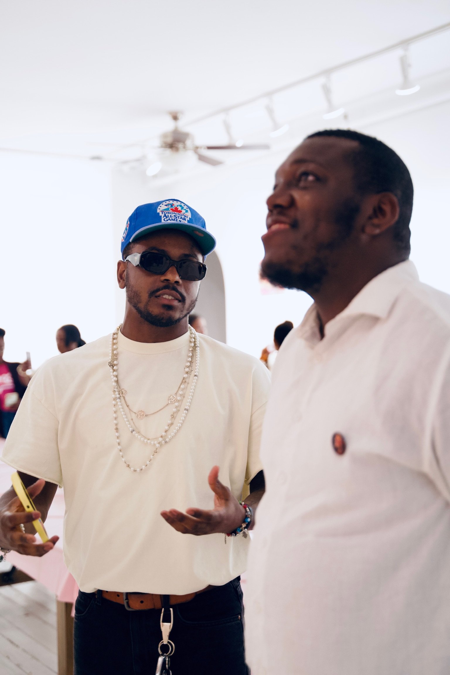 Two men engaged in conversation in a bright indoor setting, with people in the background. One man wears sunglasses, a blue cap with a baseball design, multiple pearl necklaces, and a white shirt. The other man, in a white shirt, is partially out of 