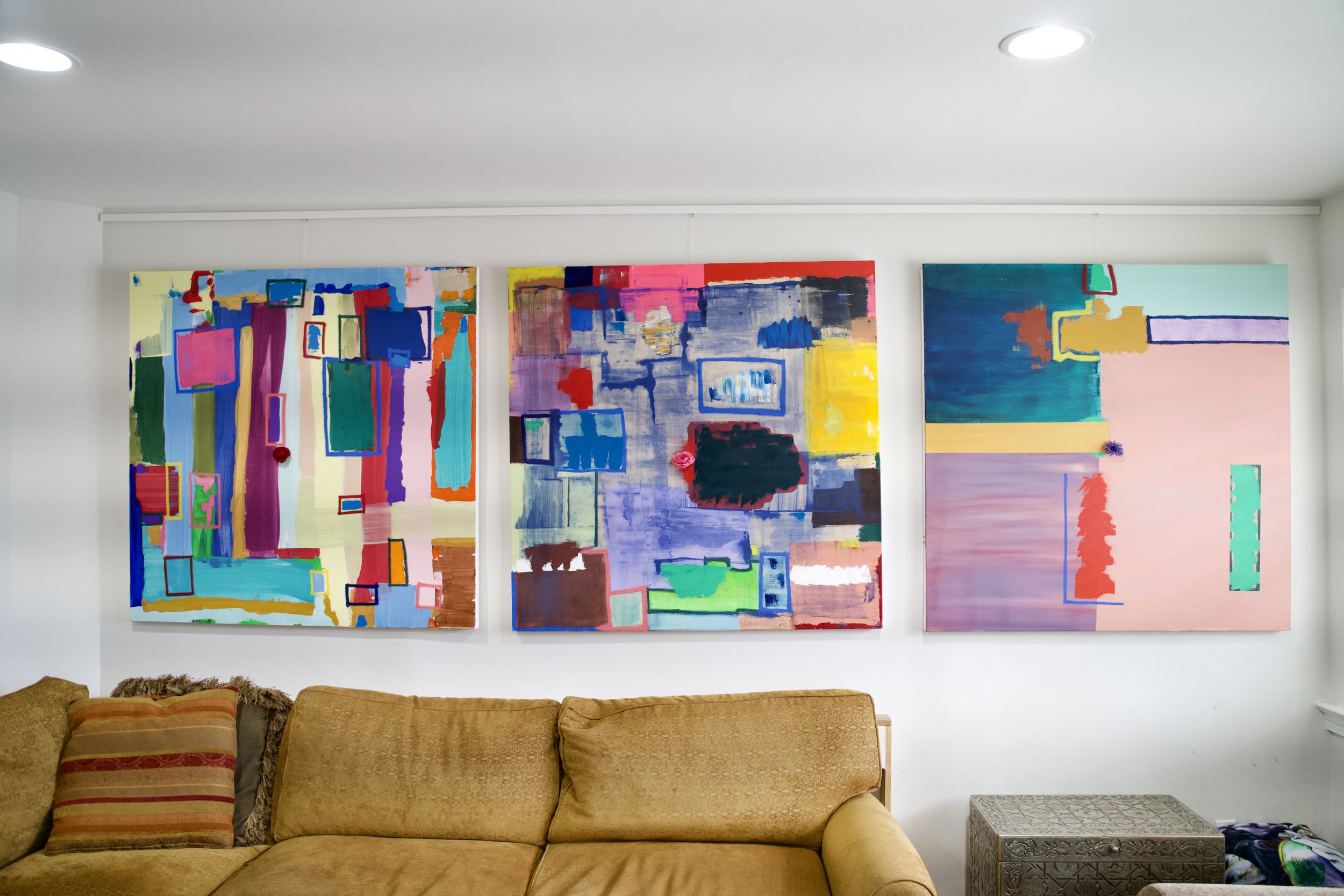 Three abstract colorful paintings hanging on a white wall in a living room with a beige sofa below them.