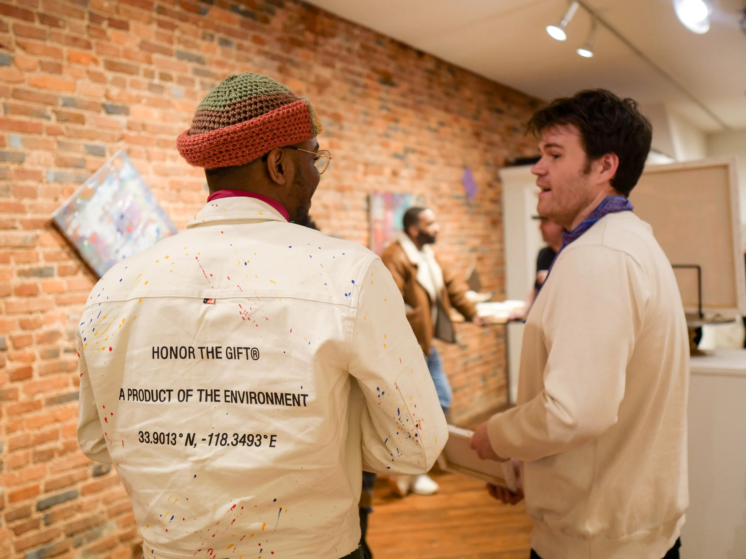 Two men talking at an art gallery. One is wearing a white jacket with colorful paint splatters and the message 'HONOR THE GIFT® A PRODUCT OF THE ENVIRONMENT' along with geographic coordinates. The other man is in a beige jacket. The gallery has a bri