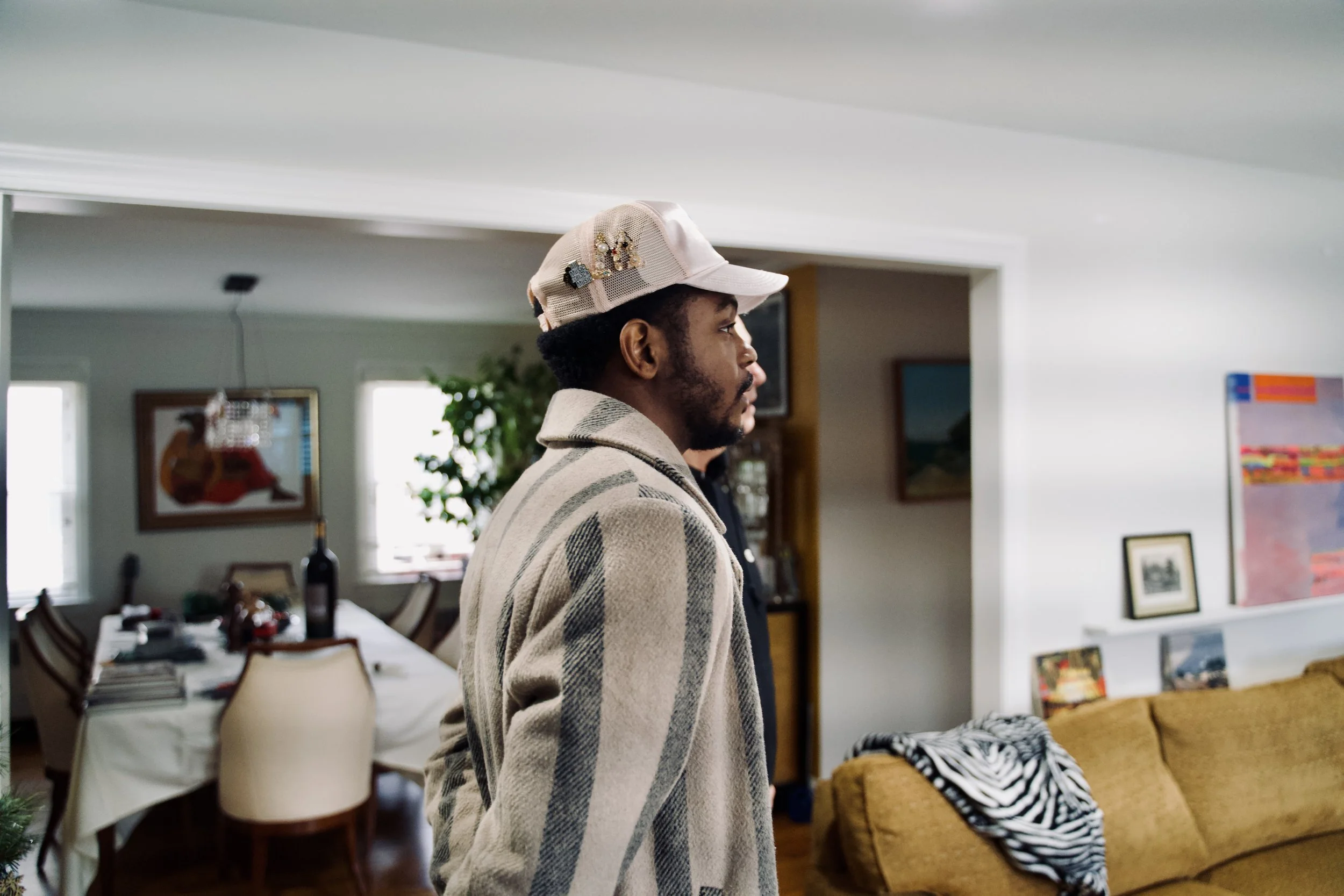A man with a beard wearing a beige checkered coat and a beige cap decorated with pins, standing indoors in a living room with some furniture and artwork in the background.