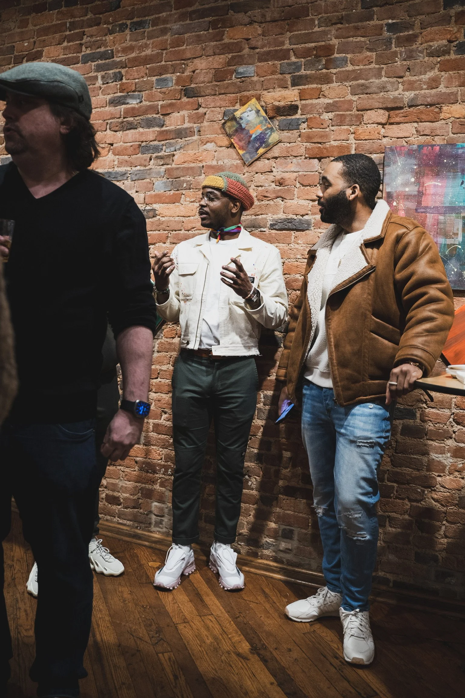 Three men standing against an exposed brick wall at an art gallery or exhibition. The man in the middle, with dark skin, wearing a colorful beanie and white jacket, is speaking. The man on the right, with dark skin, in a brown jacket, contains a bear