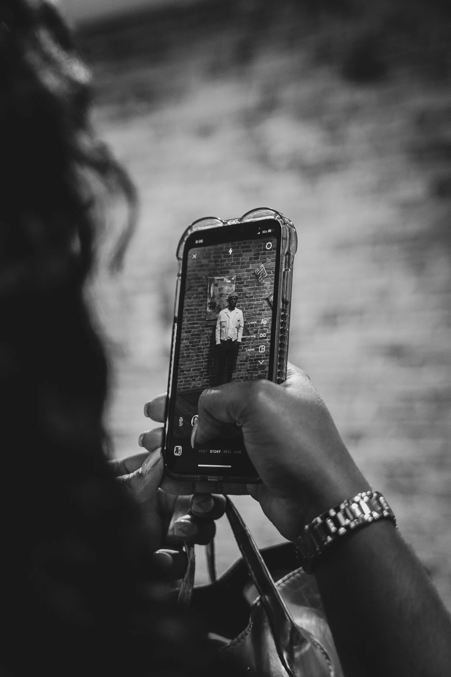 A person taking a black-and-white photo of another person standing against a brick wall with their smartphone.