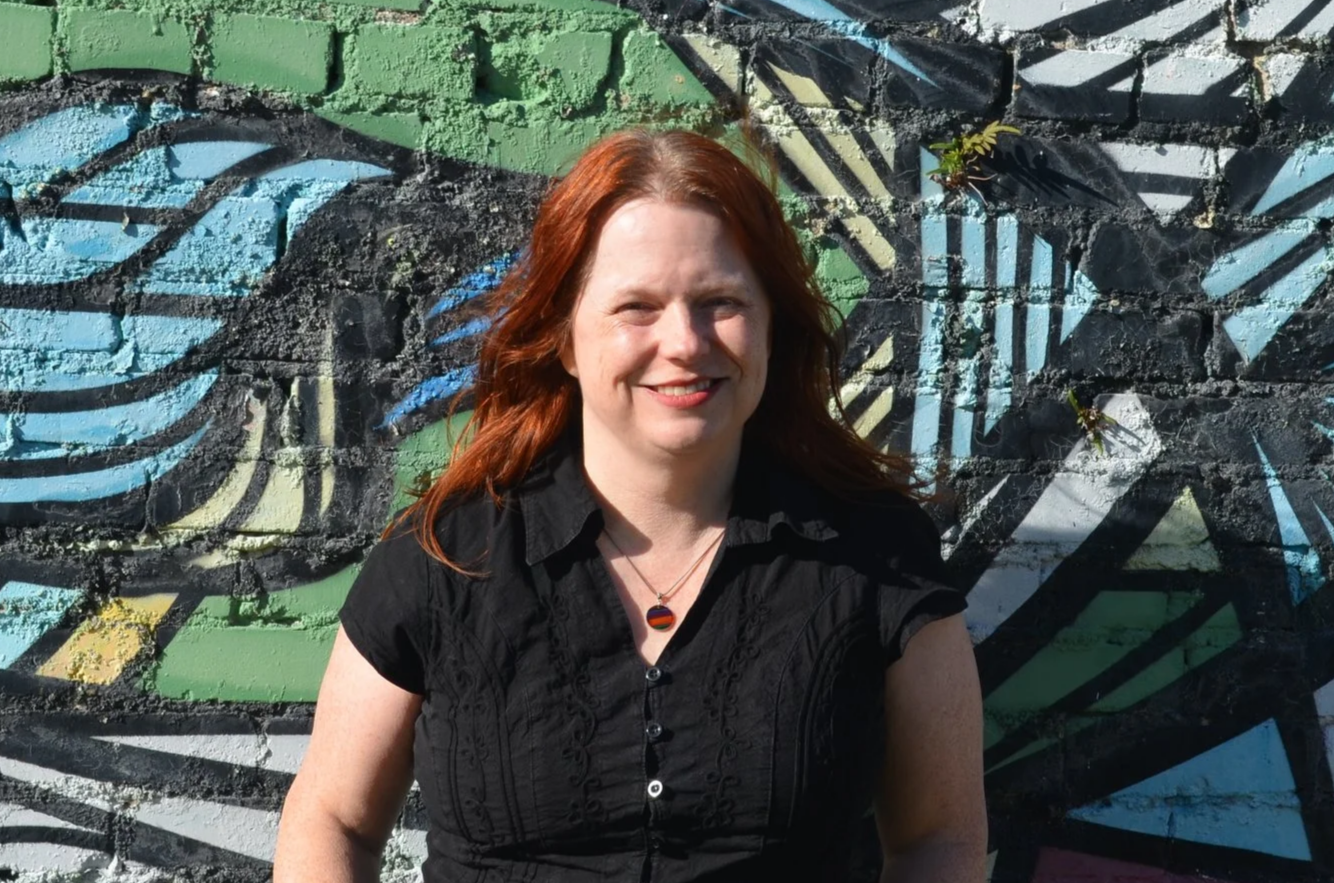 A picture of Karen Wilson, LPC-S. Karen is a Therapist at Heart of Texas Therapy Services. Karen is a woman with red hair smiling standing in front of a colorful graffiti wall.