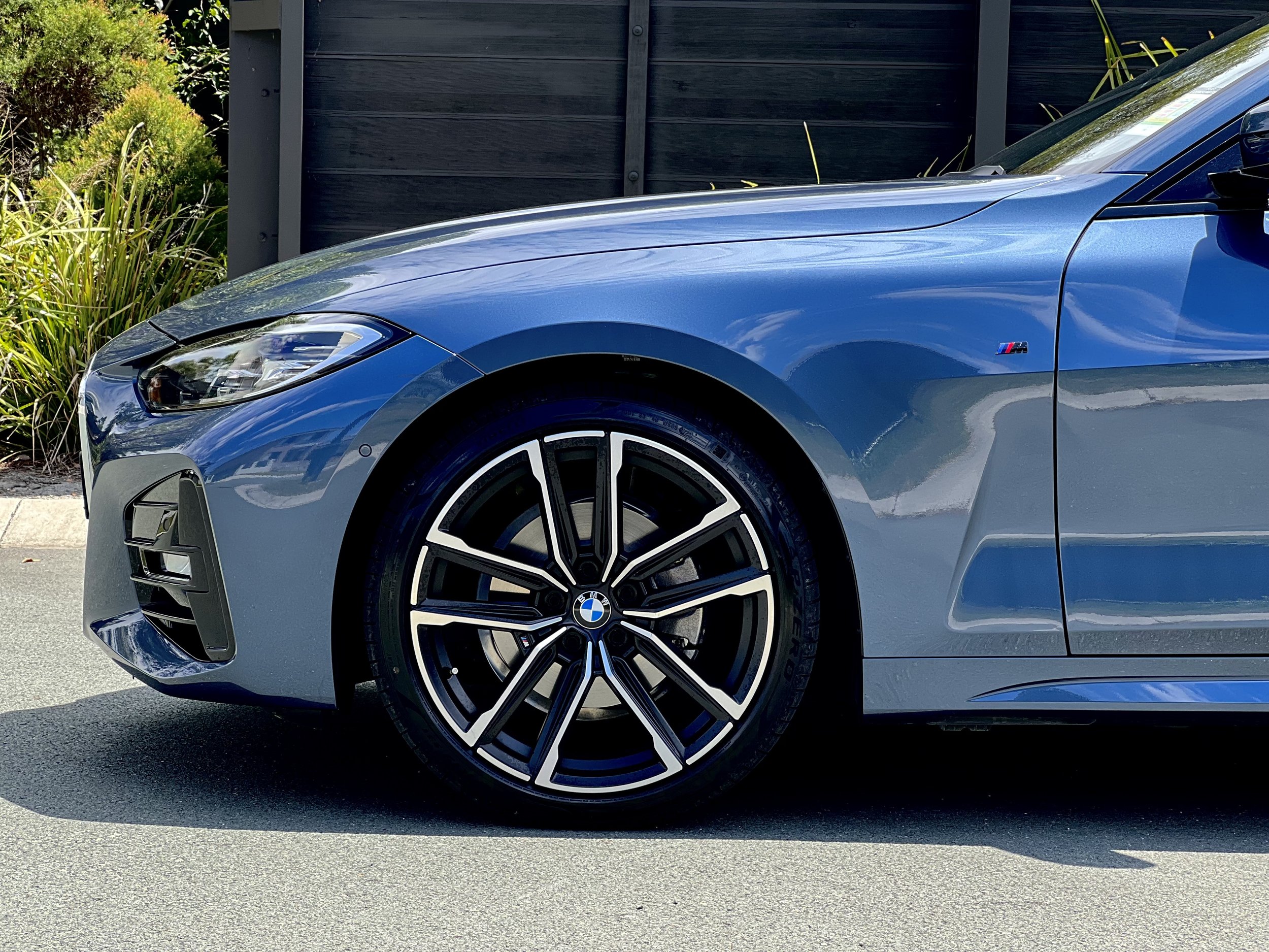 Close-up of the front section of a blue BMW car, focusing on the front wheel and headlight, with a background of greenery and a dark fence.