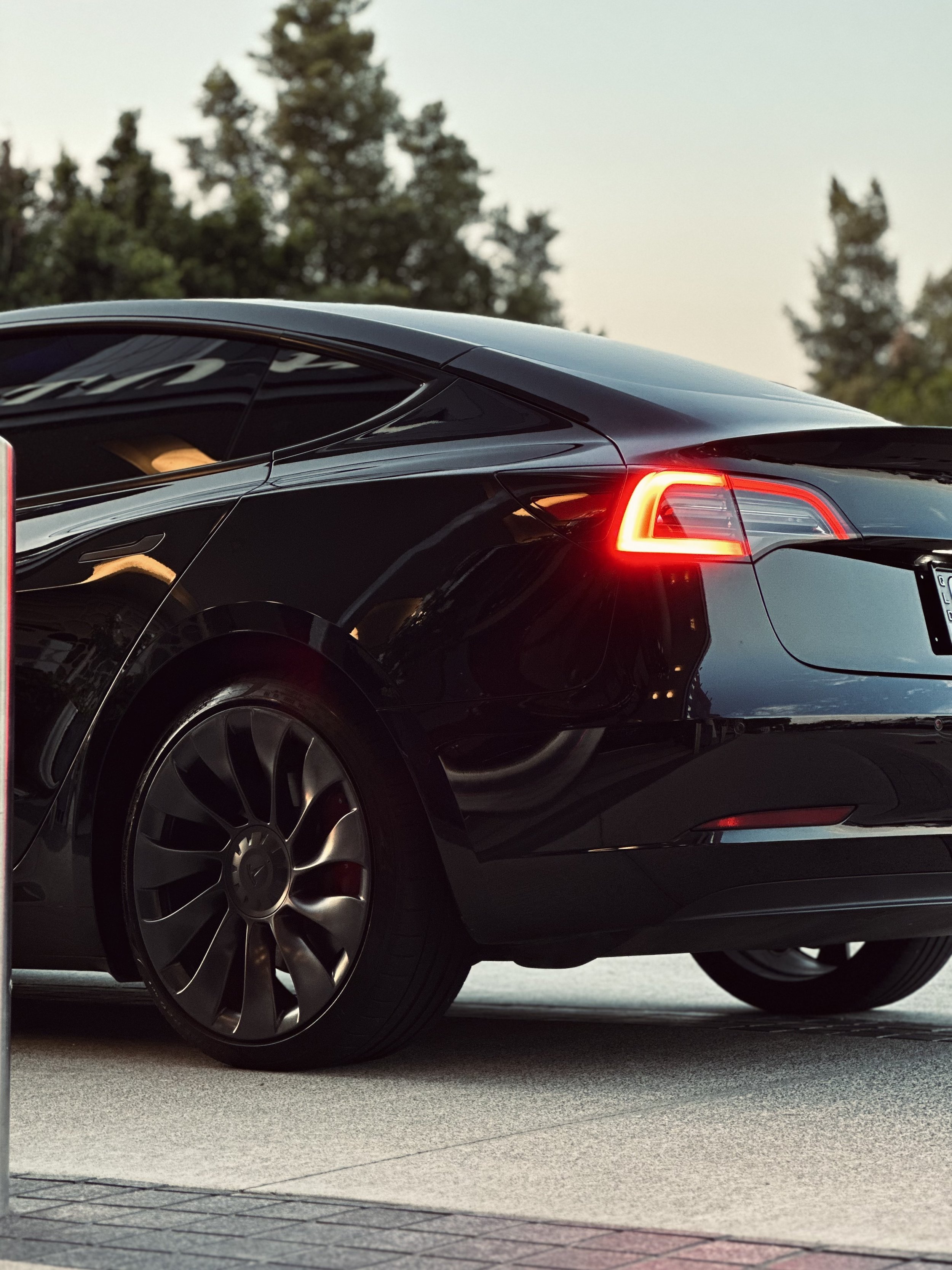 Black car with illuminated taillight, parked on a street with trees in the background.