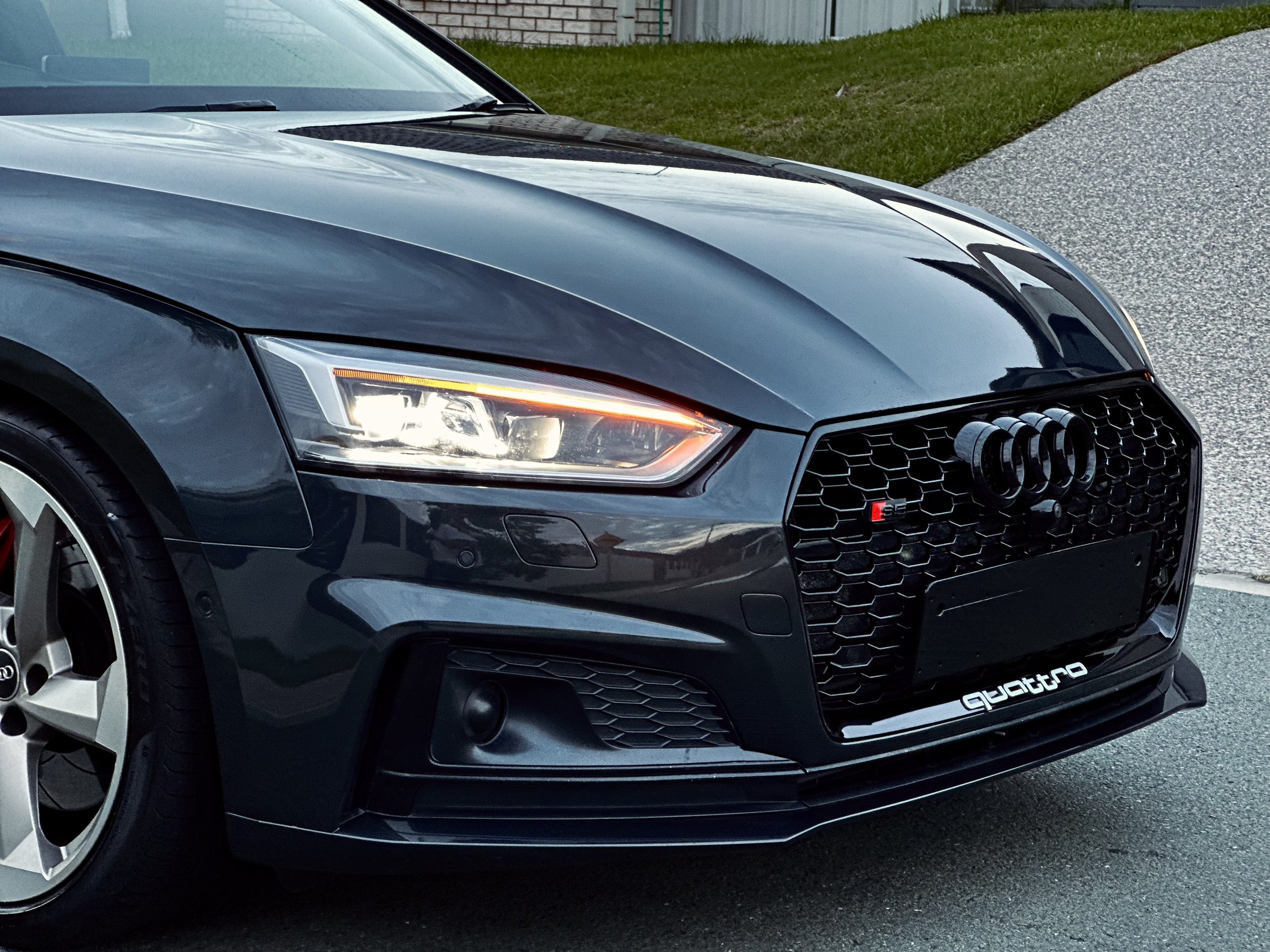 Close-up of a dark gray Audi car front, featuring LED headlights, a honeycomb grille with logo, and "quattro" badge on a road.