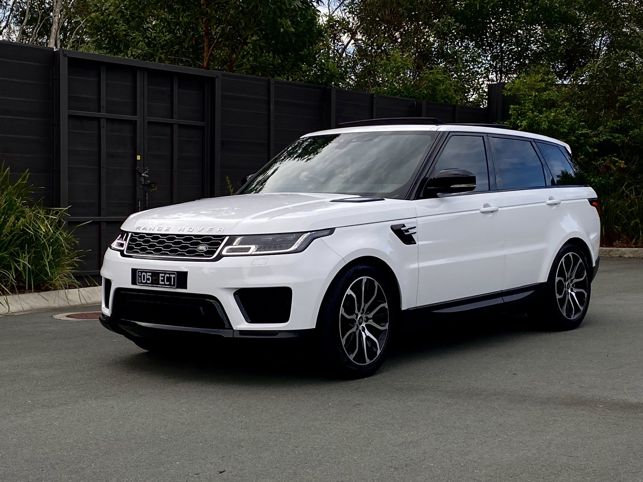 White Range Rover Sport parked near a black fence with greenery in the background.