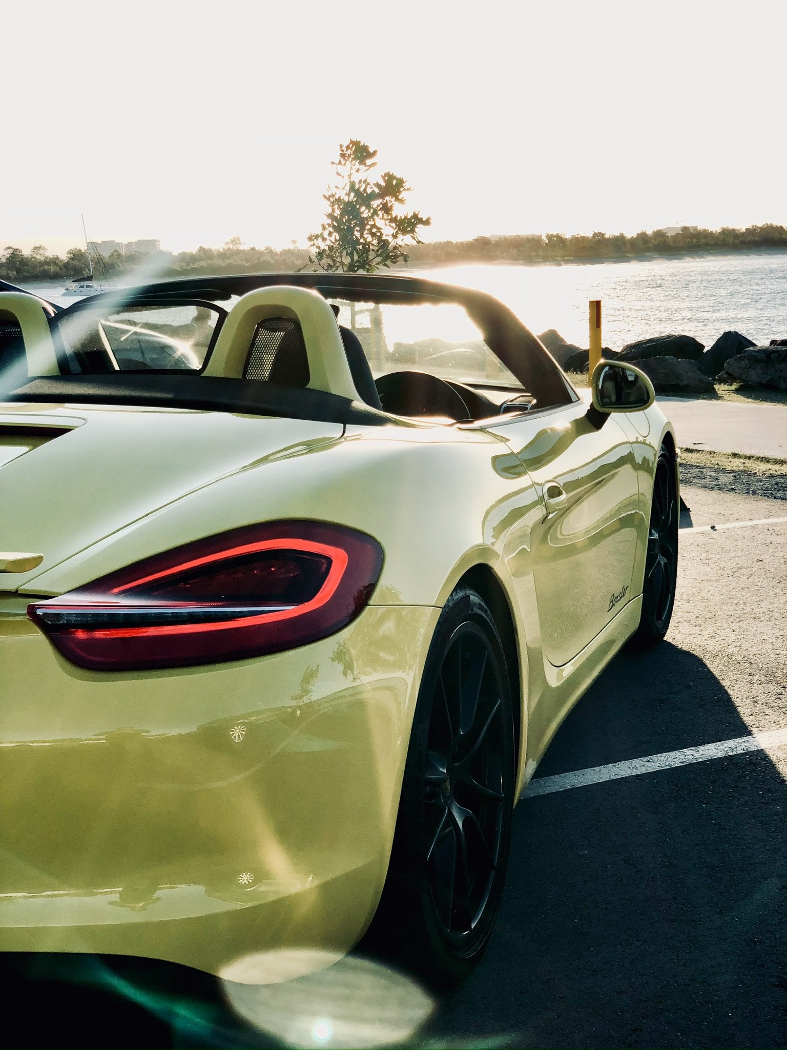 Yellow convertible sports car parked near a waterfront during sunset, with reflections on the car and serene water backdrop.