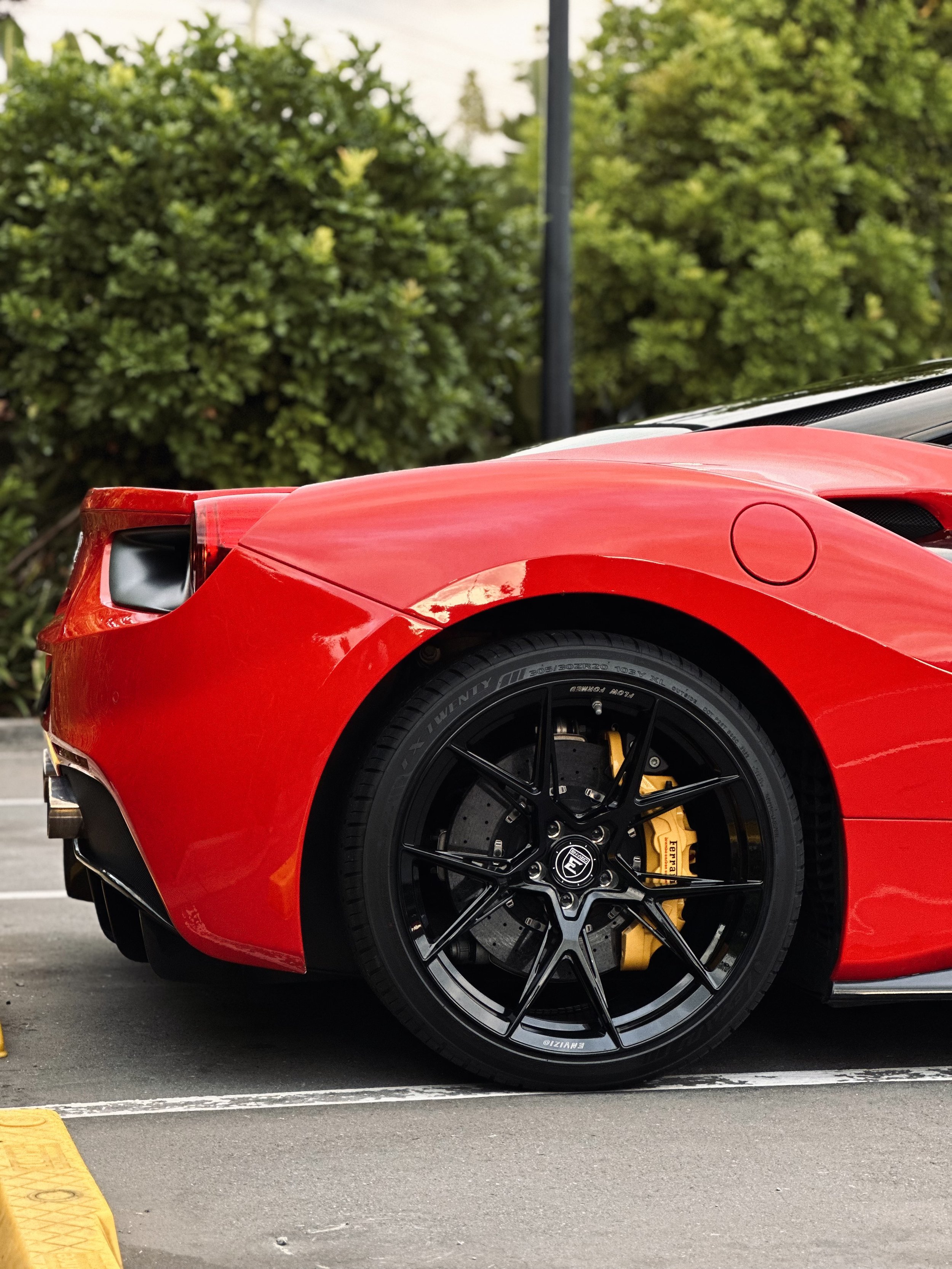 Rear side view of a red sports car with black alloy wheels and visible brake caliper, parked near green foliage.