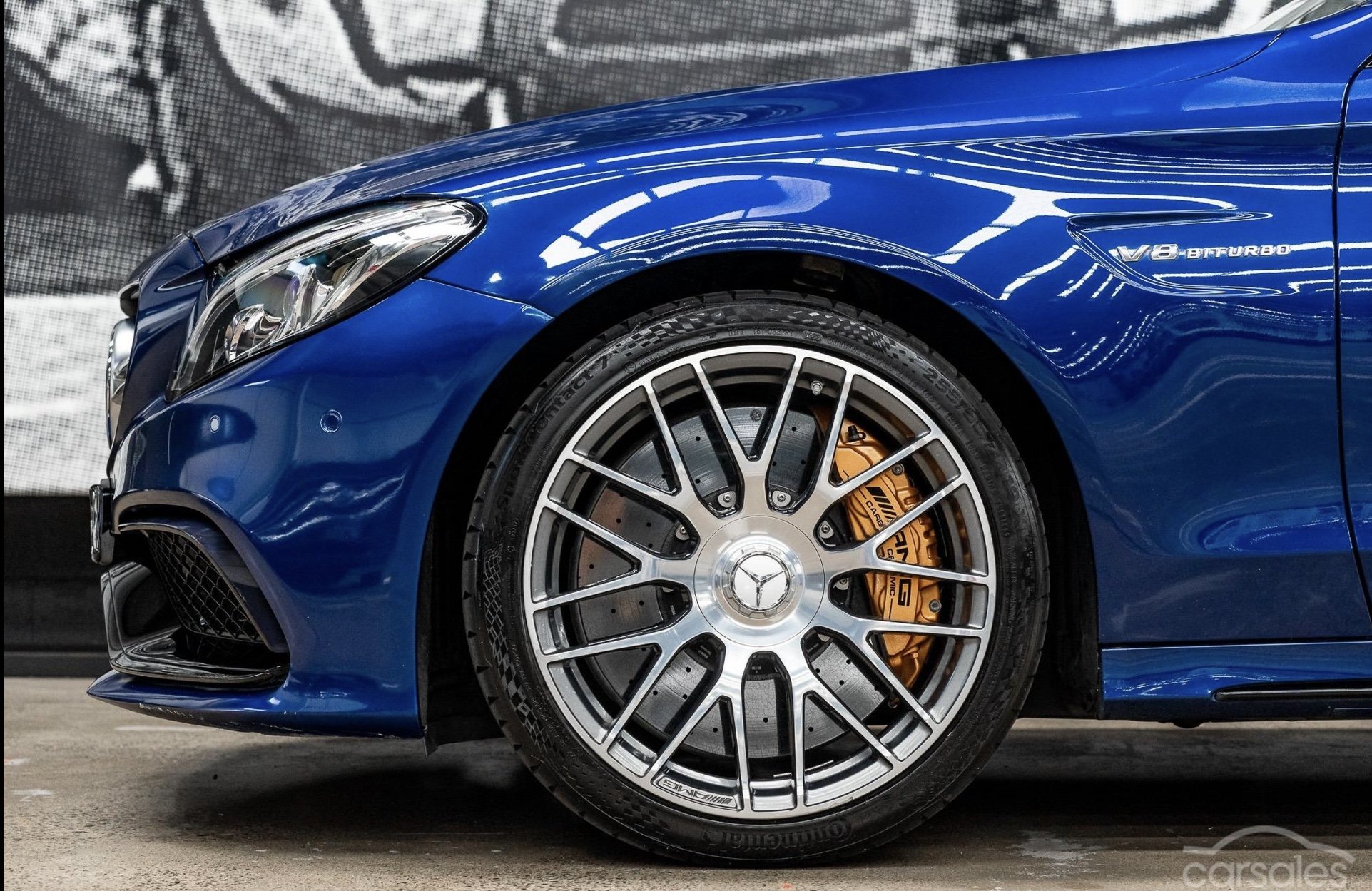 Close-up of a blue car's front wheel showing a V8 BITURBO badge, alloy wheel with logo, and brake caliper.
