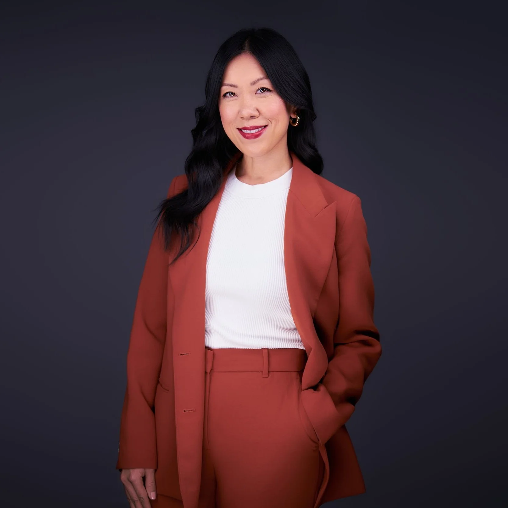 Professional woman with long black hair, wearing an orange suit and white blouse, smiling against a dark gray background.